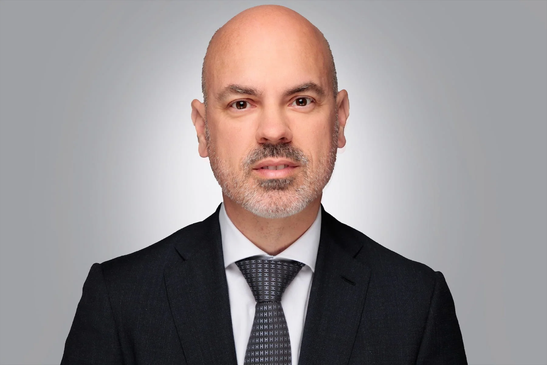 Porträt eines Mannes in formeller Kleidung mit Blond- und Grauhaar, er trägt einen schwarzen Anzug, weißes Hemd und eine schwarze Krawatte, gegen grauen Hintergrund.