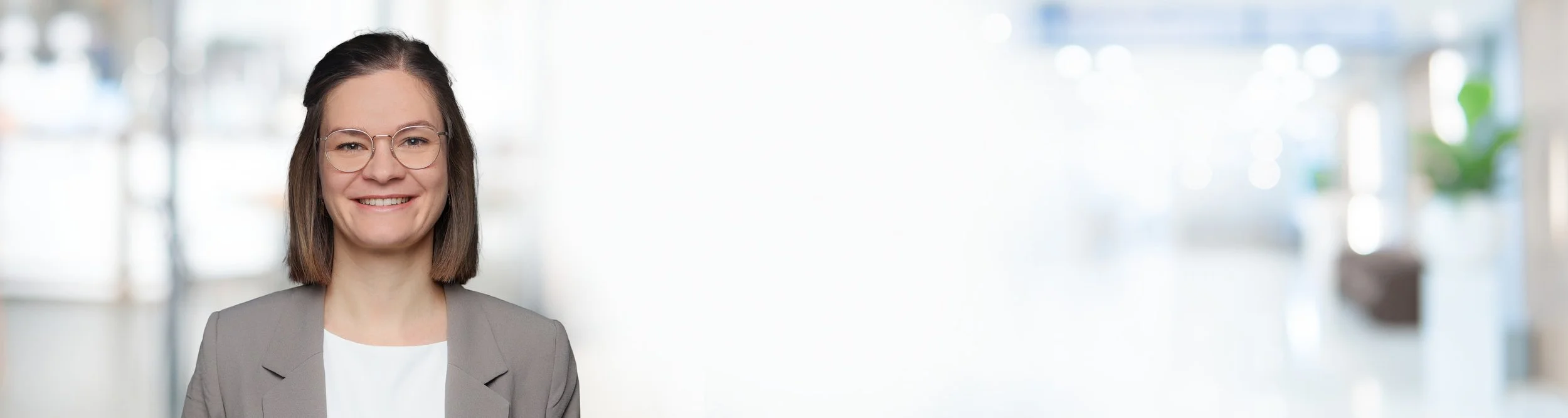 Eine lachende Frau mit Brille, grauer Blazer und weißem Hemd vor einem hellen, unscharfen Hintergrund in einem Büro.