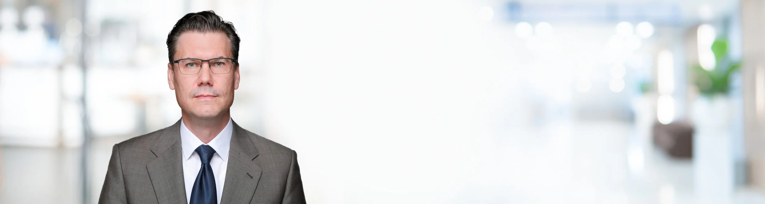 Geschäftsmann in Brille und Anzug steht vor einem hellen, unscharfen Bürohintergrund.