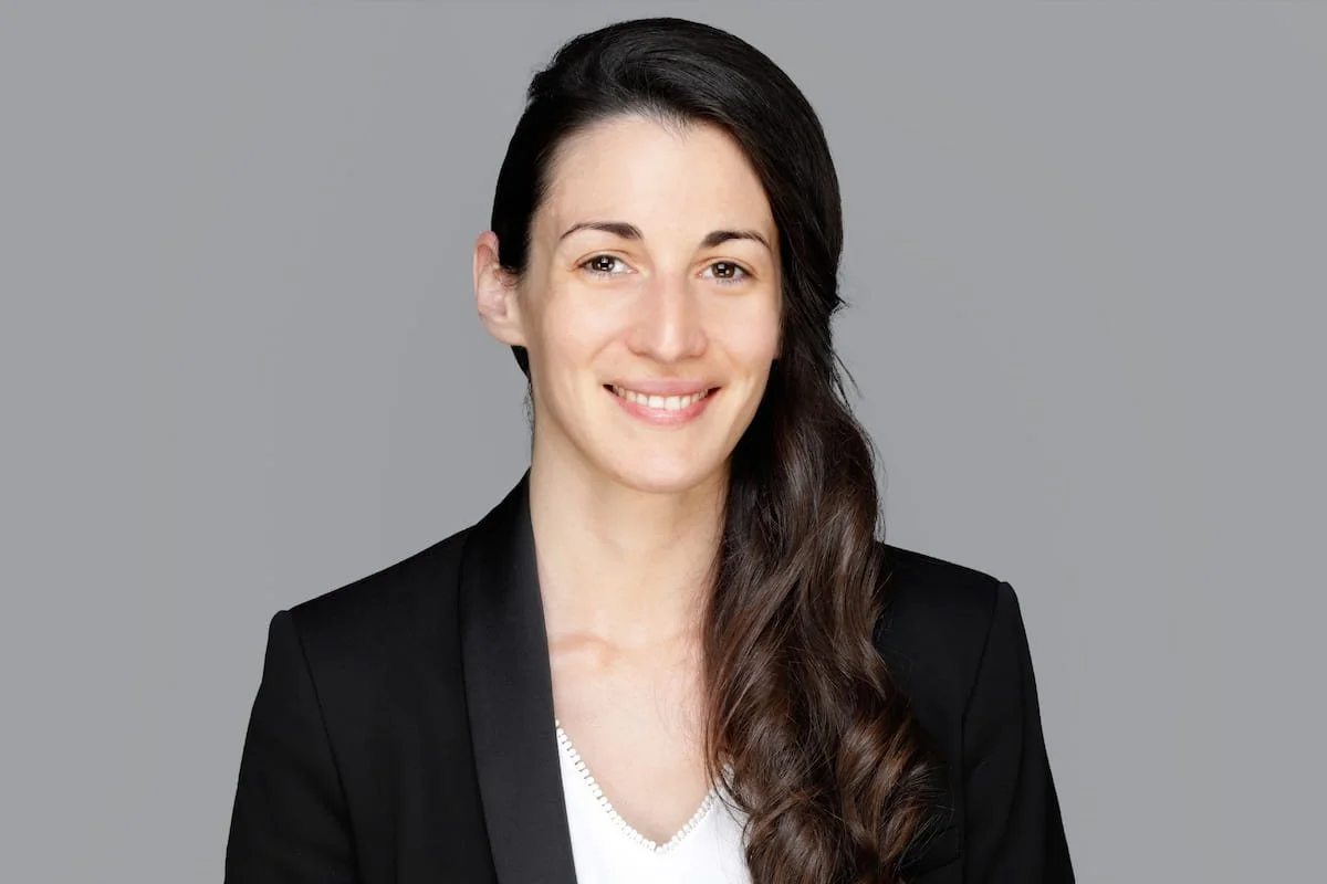 Portrait einer Frau mit langem, braunem, gewelltem Haar, schwarzer Jacke, weißem Top, auf grauem Hintergrund, lächelnd.