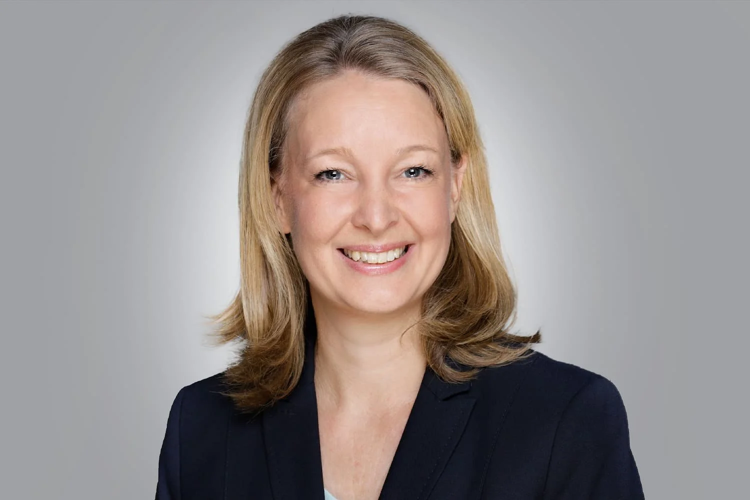 Porträt einer Frau mit braunen haaren, lächelnd, im dunklen Blazer vor hellgrauem Hintergrund.