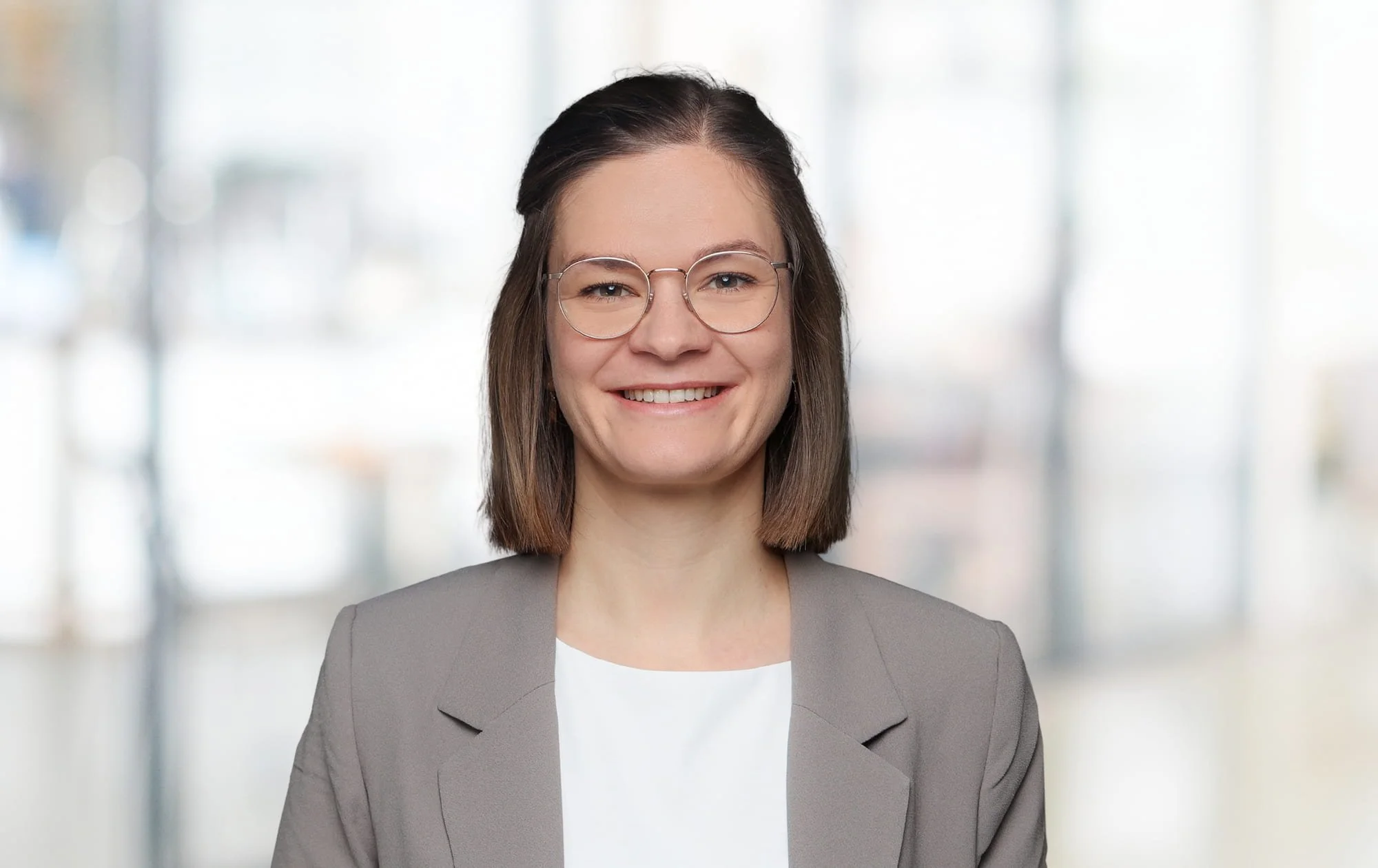 Porträt einer lächelnden Frau mit Brille in einem schicken grauen Blazer, vor einem unscharfen modernen Bürohintergrund.