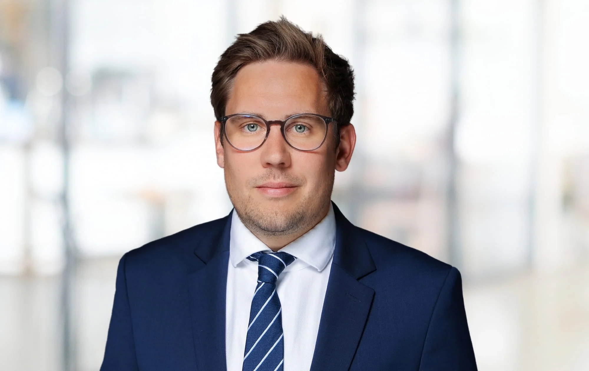 Ein Mann in einem dunklen Anzug, weißem Hemd und Lederbrille vor einem hellen, unscharfen Hintergrund.