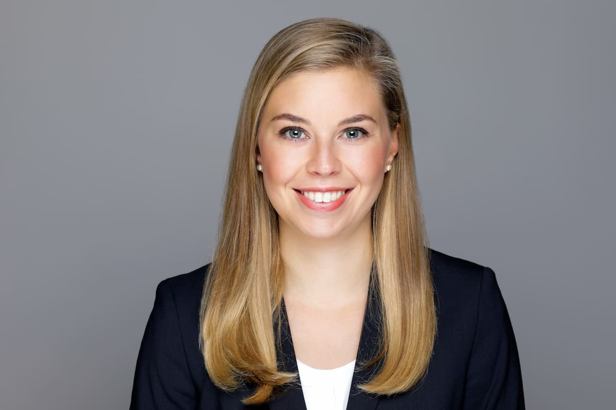 Porträt einer lächelnden jungen Frau mit langen, glatten, blonden Haaren, in formeller Kleidung vor einem grauen Hintergrund.