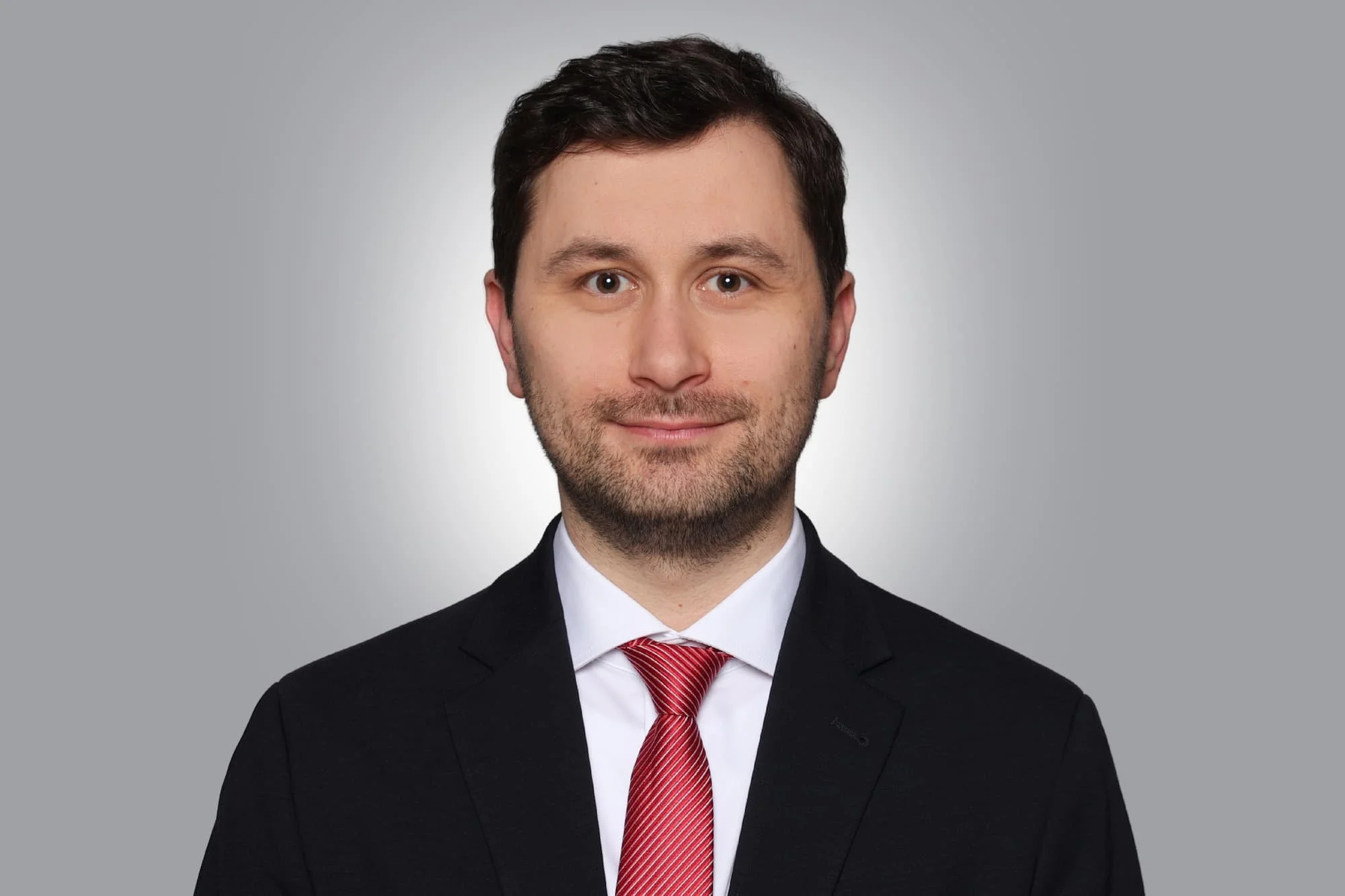Porträt eines Mannes, der freundlich schaut, in formeller Kleidung mit einem schwarzen Anzug, weißem Hemd und roter Krawatte vor grauem Hintergrund.