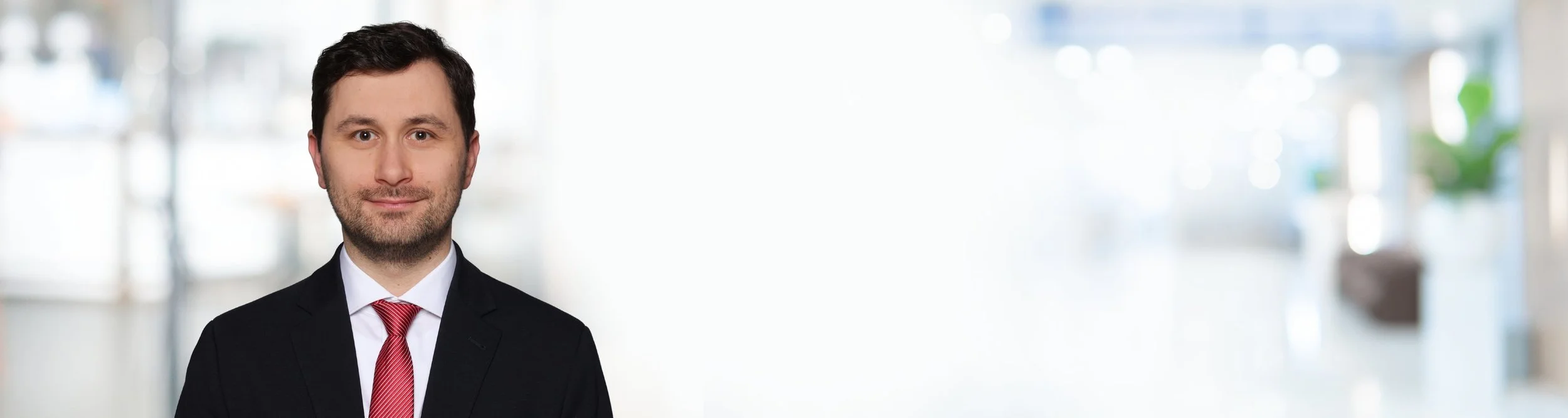 Ein Mann in einem schwarzen Anzug mit roter Krawatte, vor einem verschwommenen hellen Hintergrund.