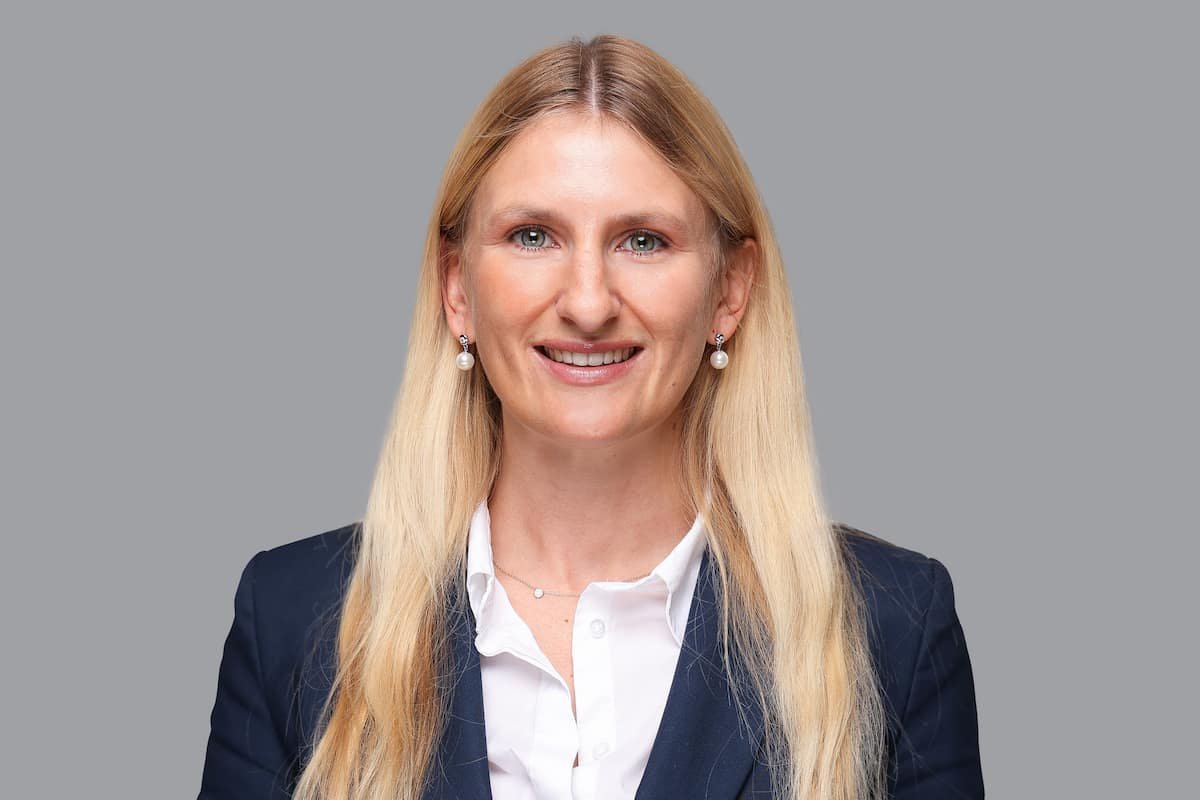 Professionelles Portrait einer lächelnden Frau mit langen blonden Haaren, weißem Hemd, dunklem Blazer, Perlenohrringen und einer zarten Halskette vor grauem Hintergrund.
