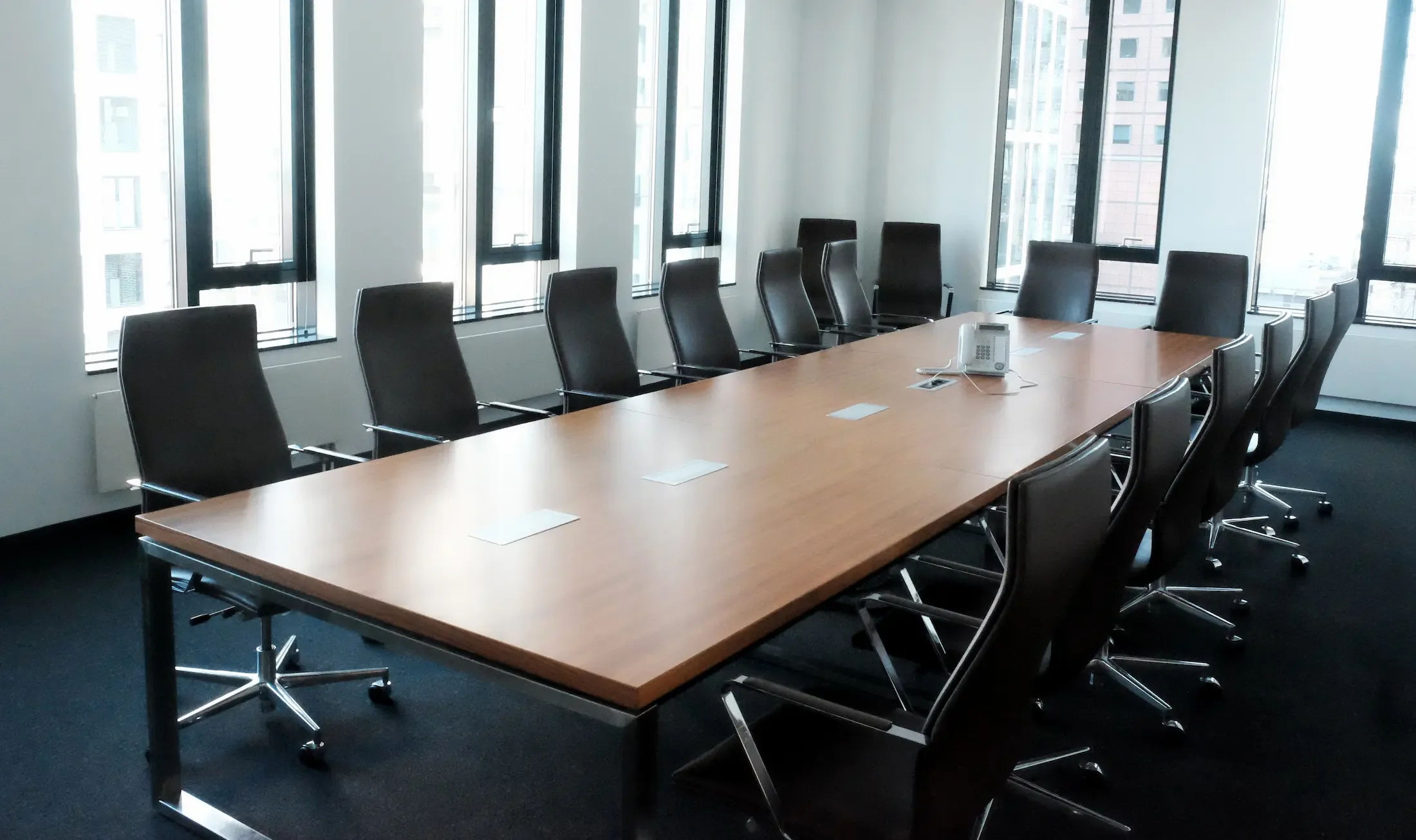Leerer Konferenzraum mit großem Holztisch, schwarzen Bürostühlen, einem Telefon und einigen Steckdosen, umgeben von großen Fenstern mit Stadtblick.