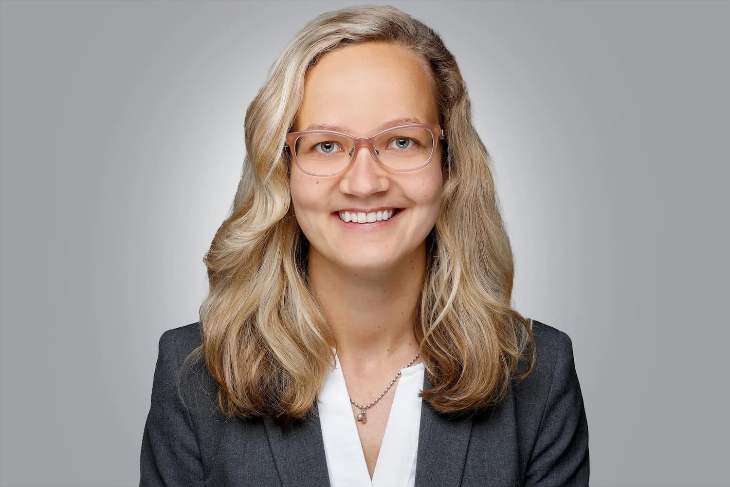 Porträt einer blonden Frau mit transparenter Brille, die freundlich lächelt, im grauen Anzug.