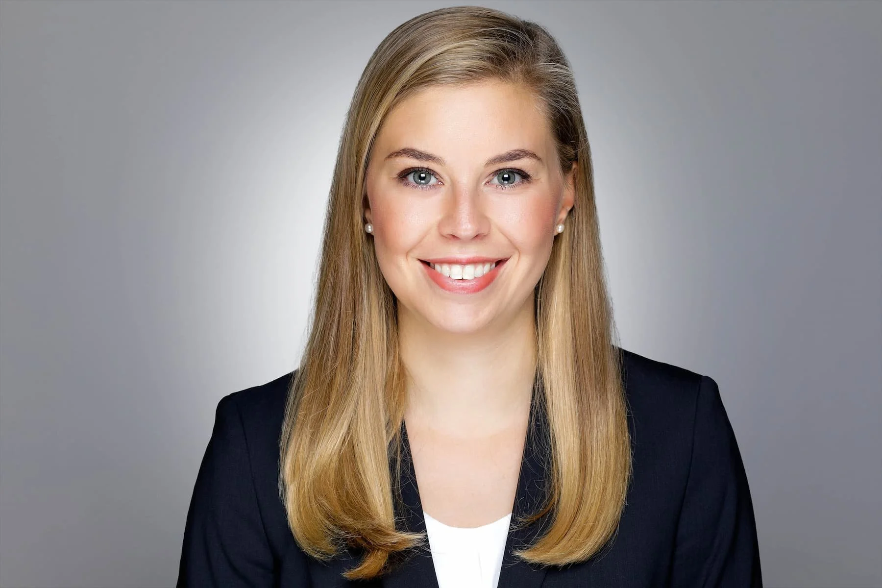Porträt einer lächelnden jungen Frau mit langen, glatten, blonden Haaren, in formeller Kleidung vor einem grauen Hintergrund.