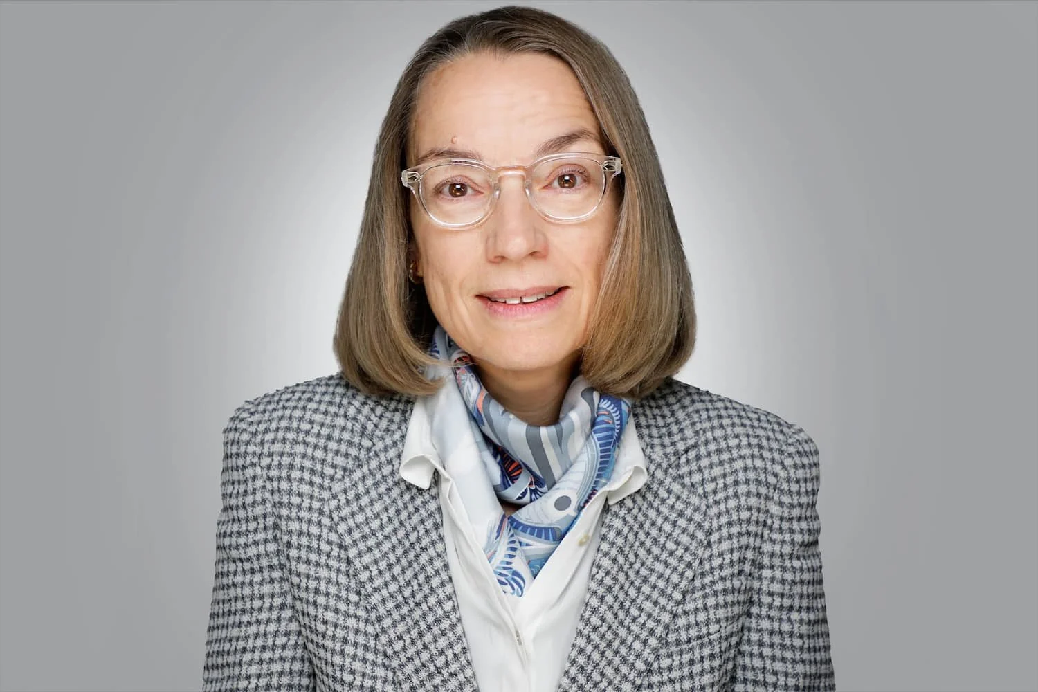 Porträt einer Anwältin mit schulterlangem Haar und transparenter Brille, grauem kariertem Blazer, weißer Bluse und gemustertem Seidentuch.