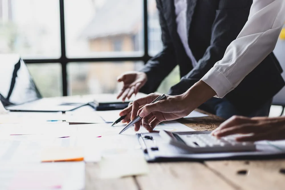Zwei Geschäftsleute arbeiten an einem Tisch mit Dokumenten, einem Laptop, einem Laptop, einem Notizbuch und einem Taschenrechner in einem Büro mit großen Fenstern.
