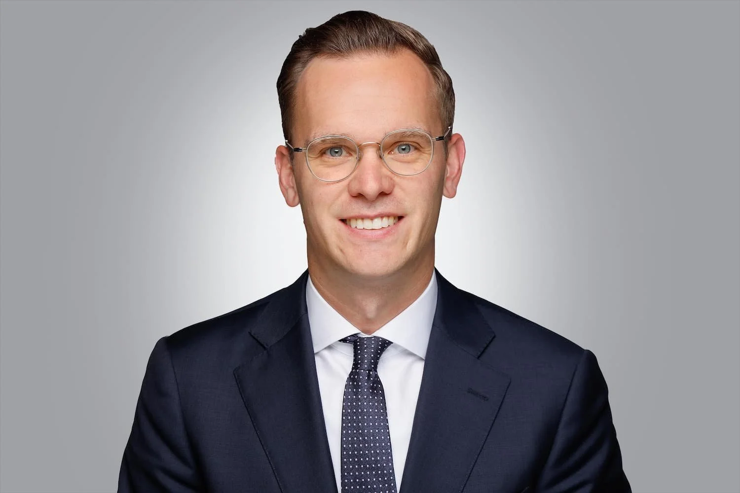 Ein Mann trägt eine dunkle Anzugjacke, ein weißes Hemd und eine dunkelblaue Krawatte mit weißen Punkten. Er trägt runde Brillen und hat kurze, braune Haare. Hintergrund ist grau.