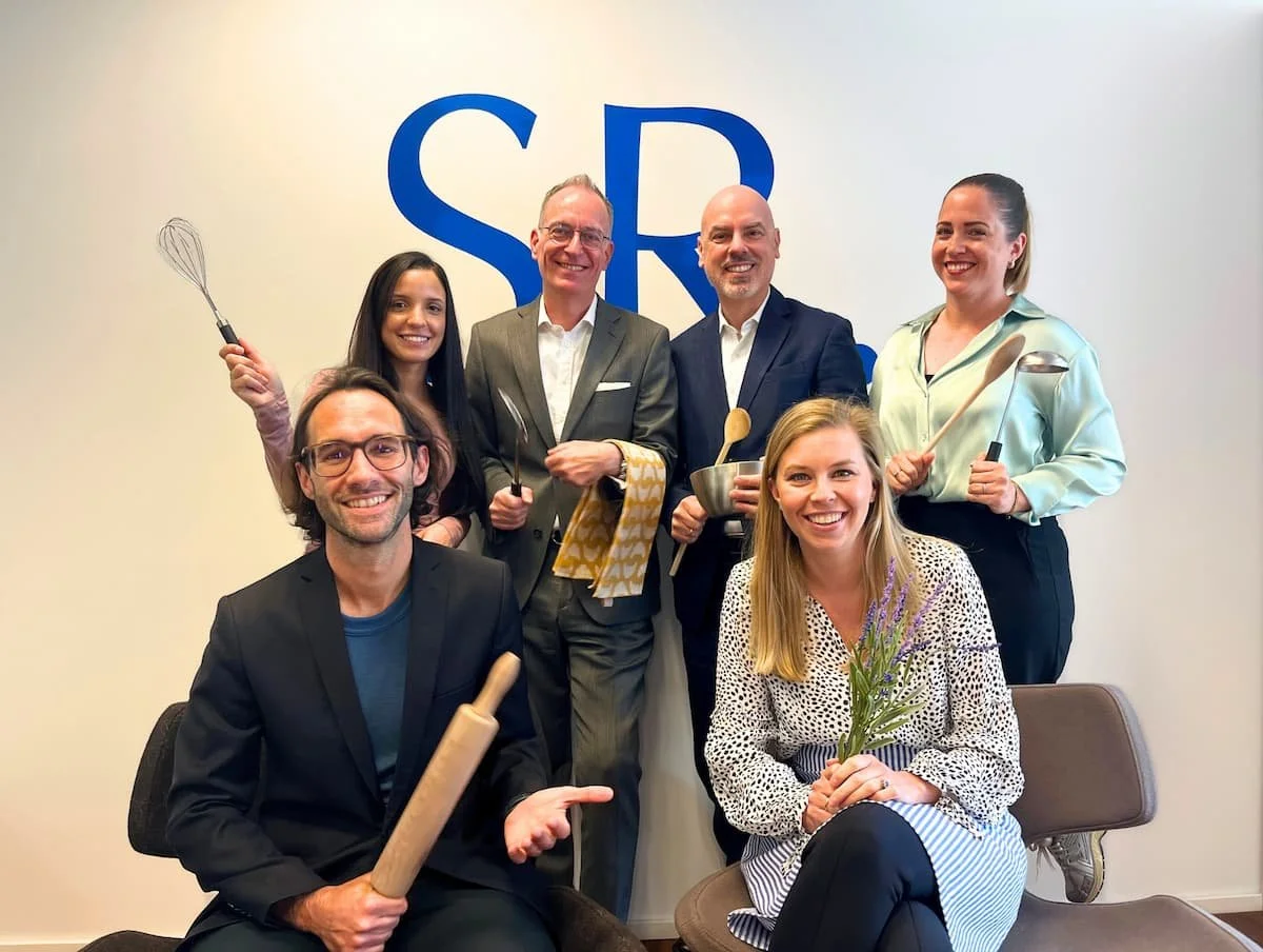 Gruppenfoto von sechs Menschen, die in einem Raum vor einer weißen Wand mit blauer Schrift stehen. Sie posieren mit Küchenutensilien und lachen. Es sind fünf Männer und eine Frau.