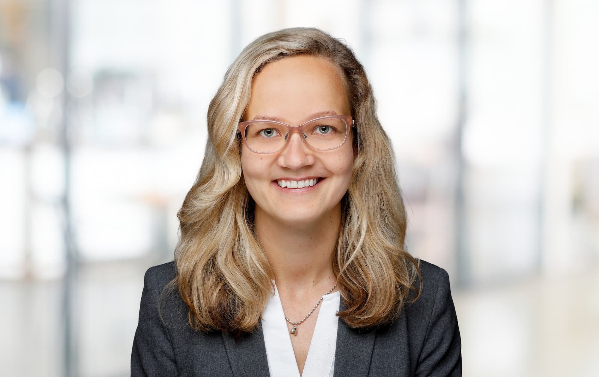 Porträt einer lächelnden Frau mit langem, gewelltem blondem Haar, Brille und dunklem Blazer vor unscharfem, hellem Hintergrund.