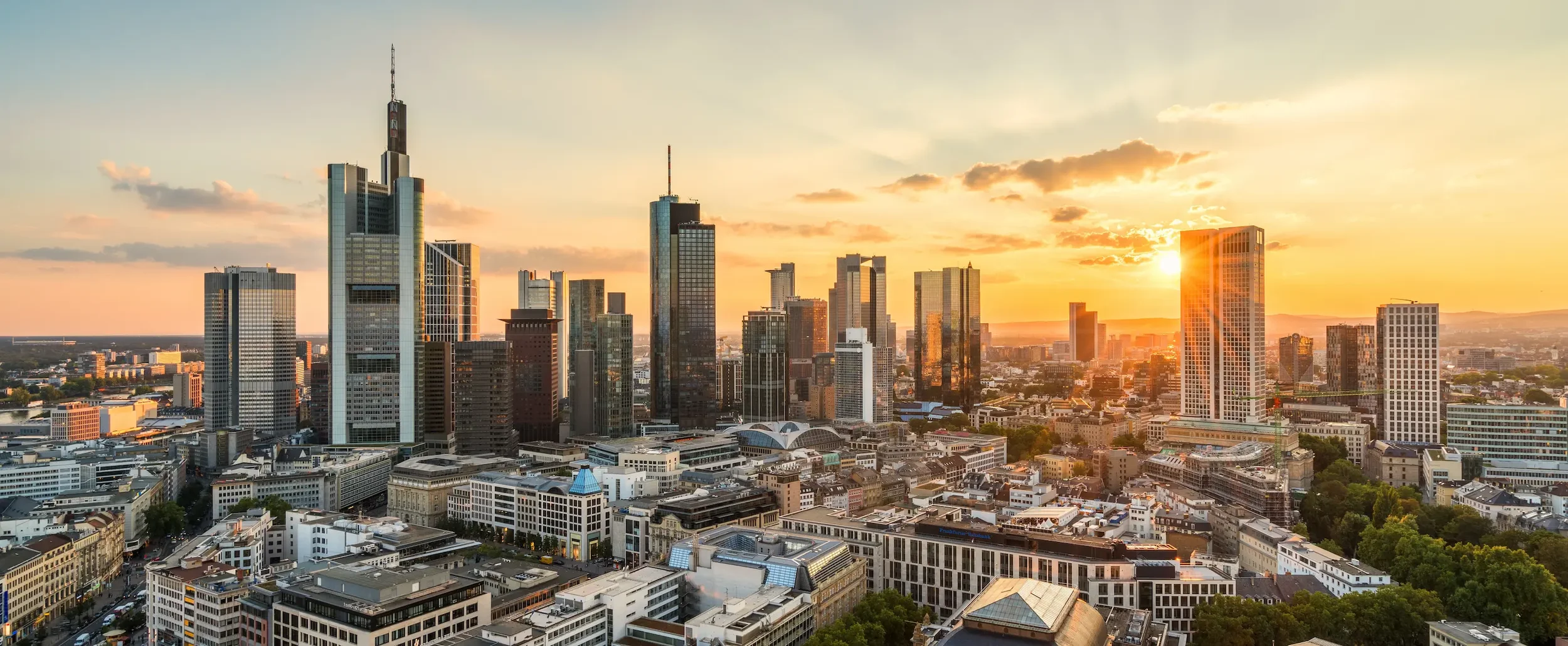 Skyline von Frankfurt am Main mit modernen Hochhäusern und Bankenviertel; das Bild ist diagonal geteilt und zeigt links eine kühlere Tagesstimmung und rechts einen warmen Sonnenaufgang über der Stadt.