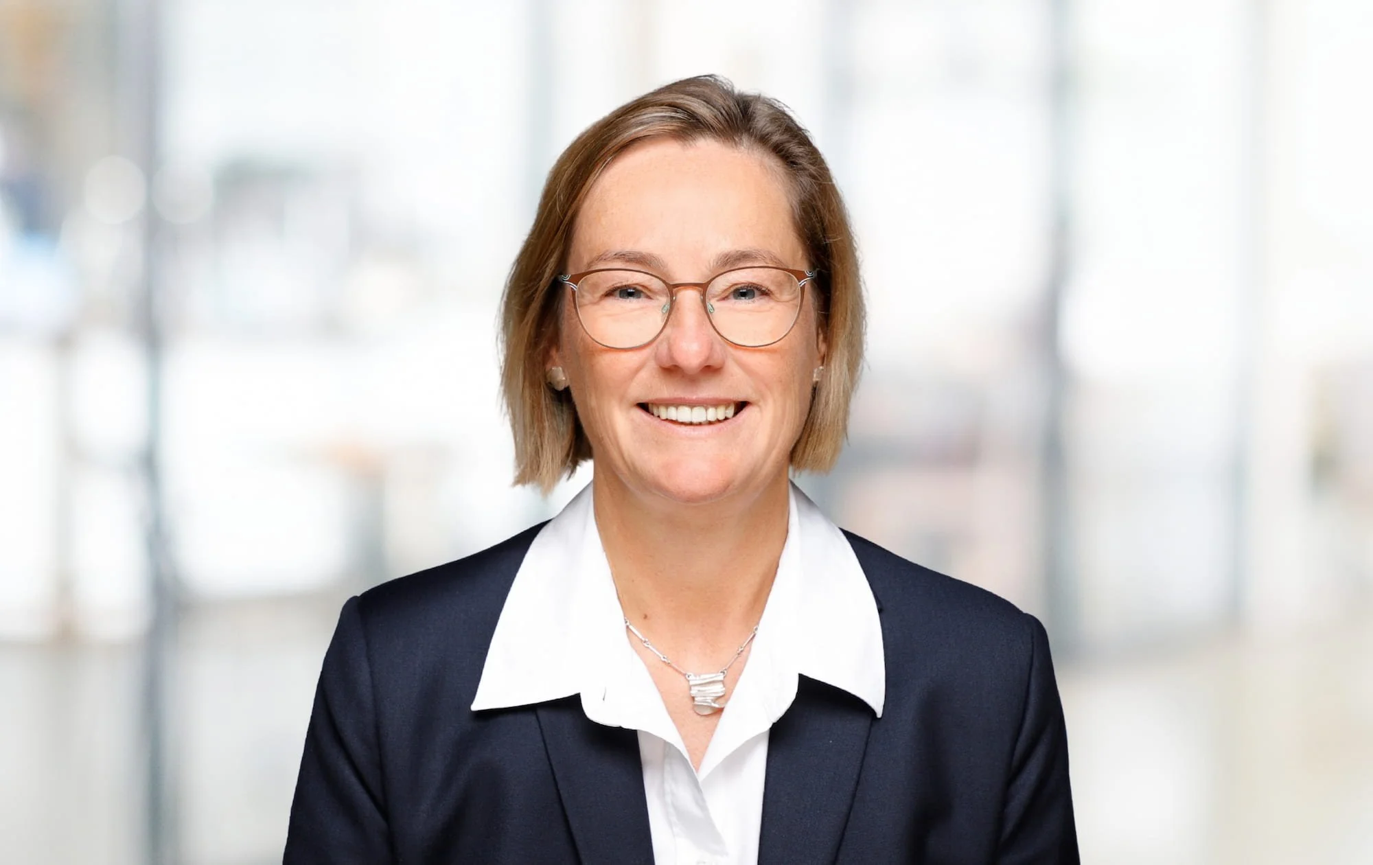 Porträt einer lächelnden Frau mit Brille in formaler Kleidung vor einem hellen, unscharfen Hintergrund.