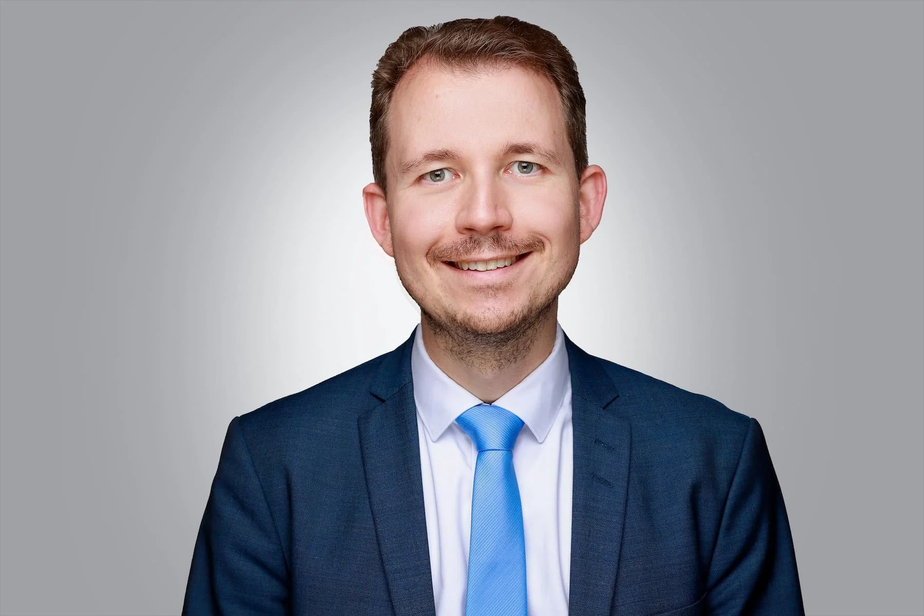Ein junger Mann trägt einen dunklen Anzug, weißes Hemd und blaue Krawatte und lächelt vor einem grauen Hintergrund.