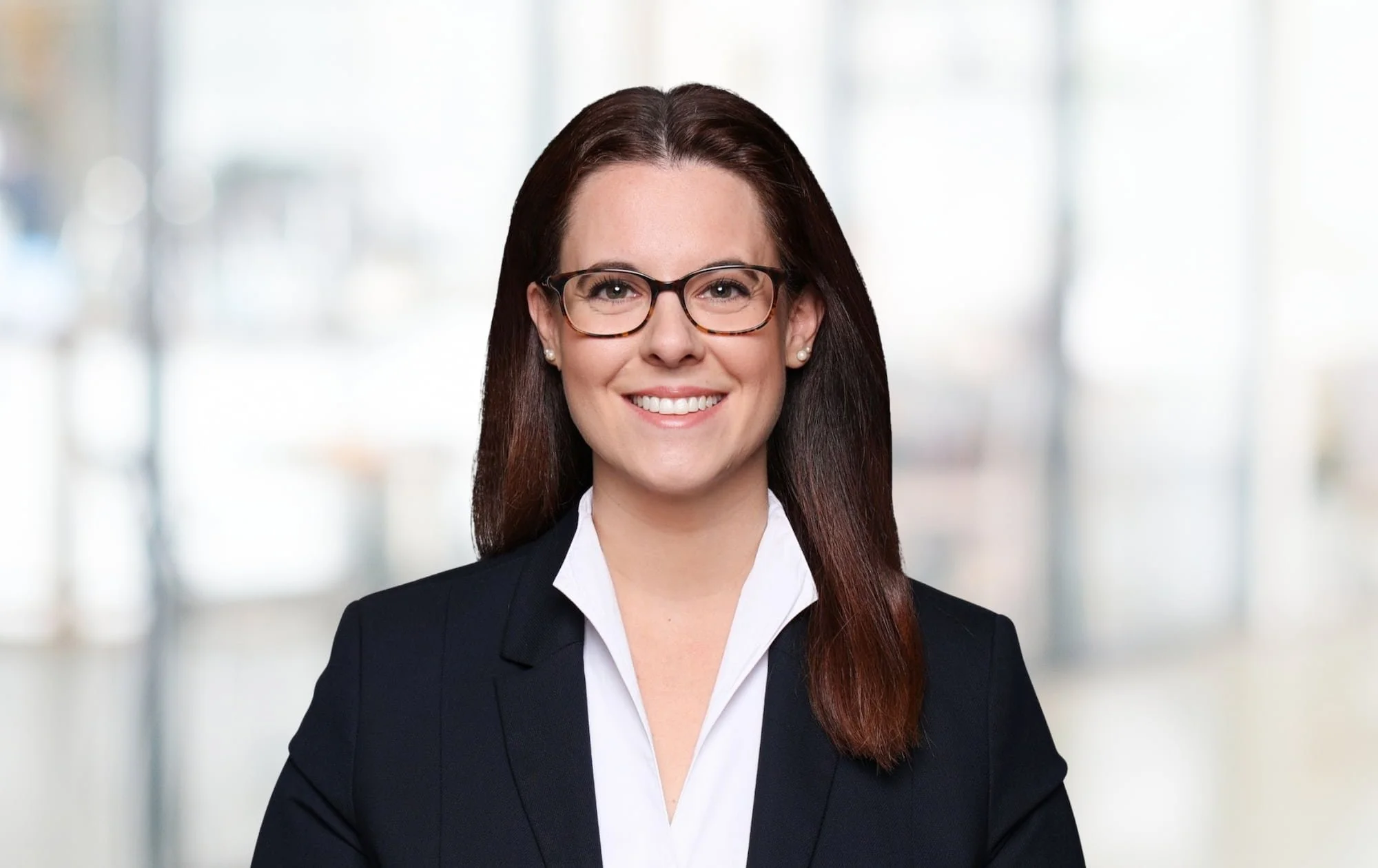 Eine lachende professionelle Frau im Business-Outfit mit Brille vor einem verschwommenen Büro-Hintergrund.