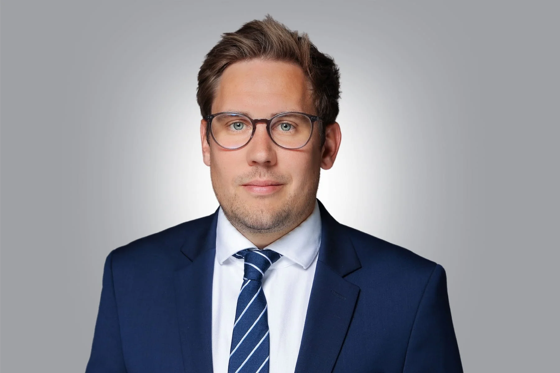 Porträt eines Mannes im Anzug mit Krawatte und Brille vor grauem Hintergrund, seriöser Business-Look.