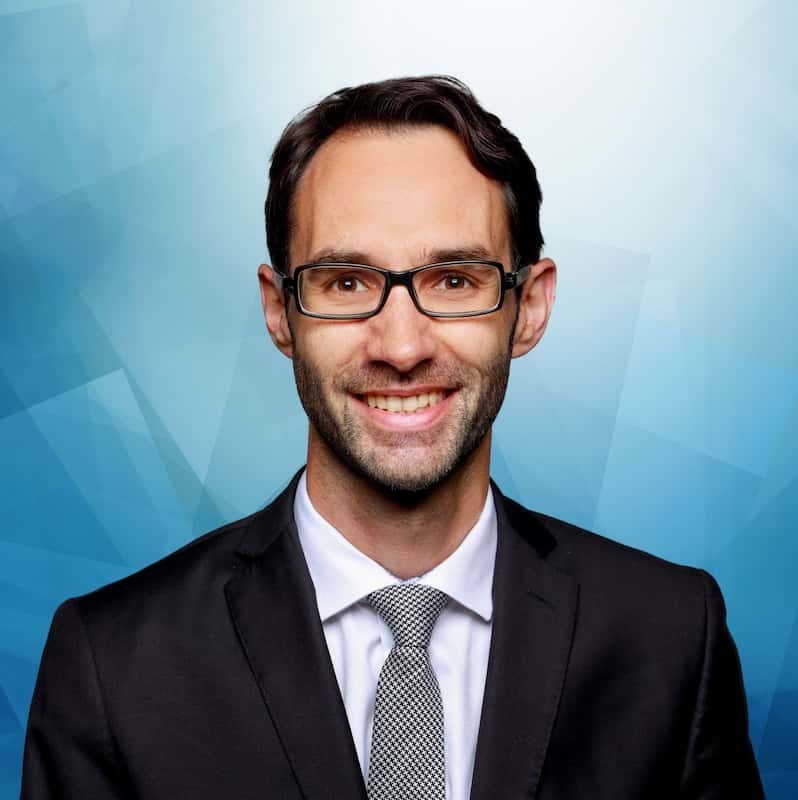 Professionell aussehender Mann mit dunklen Haaren, Brille, Anzug und Krawatte, lächelnd vor blauem geometrischem Hintergrund.