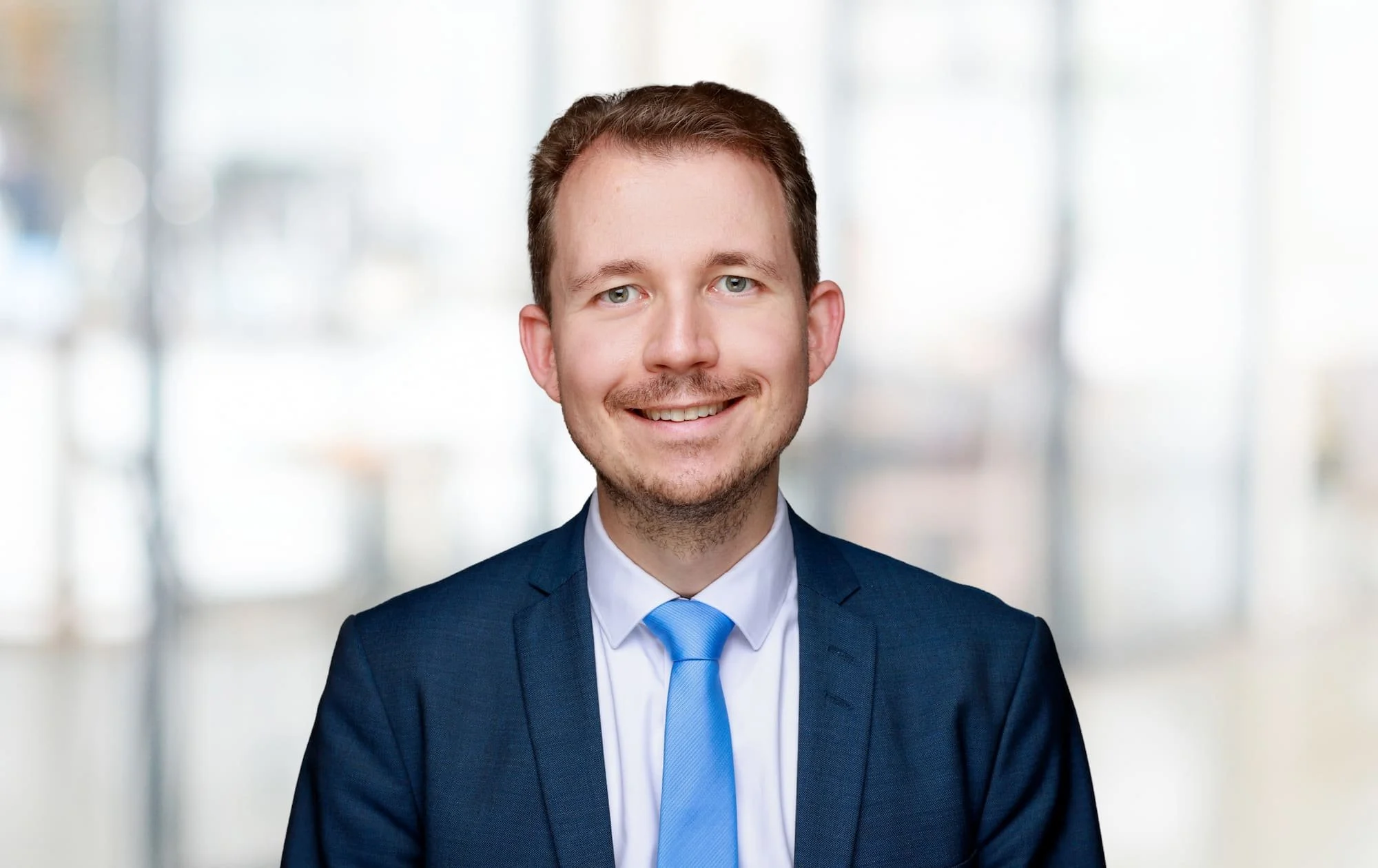 Junger Mann in dunklem Anzug mit blauer Krawatte vor hellem, unscharfem Hintergrund.