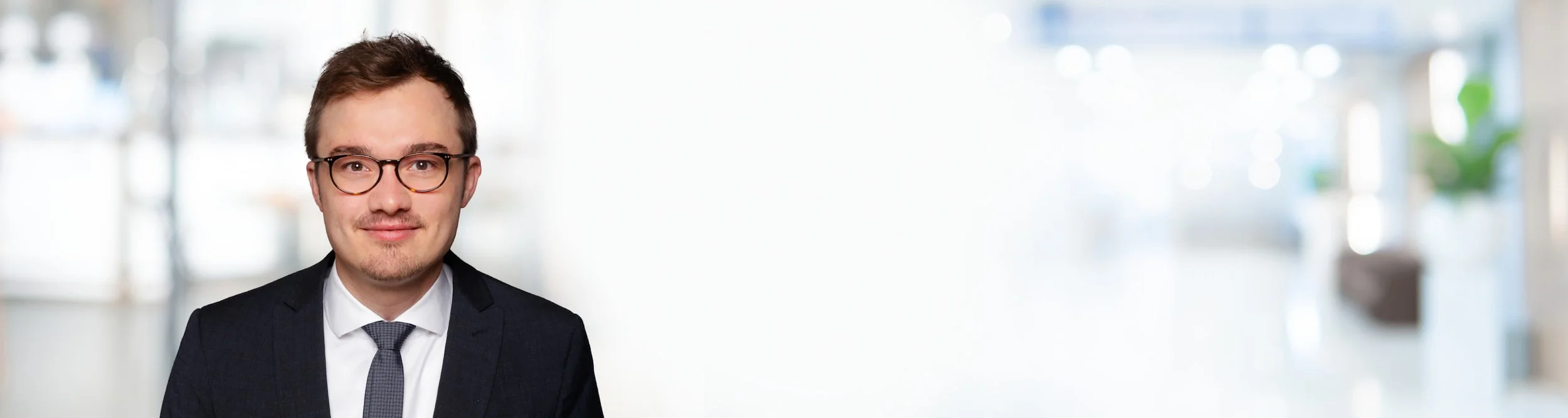 Porträt eines Mannes in Anzug und Brille vor einem hellen, modernen Büro mit großflächigem Fenster im Hintergrund