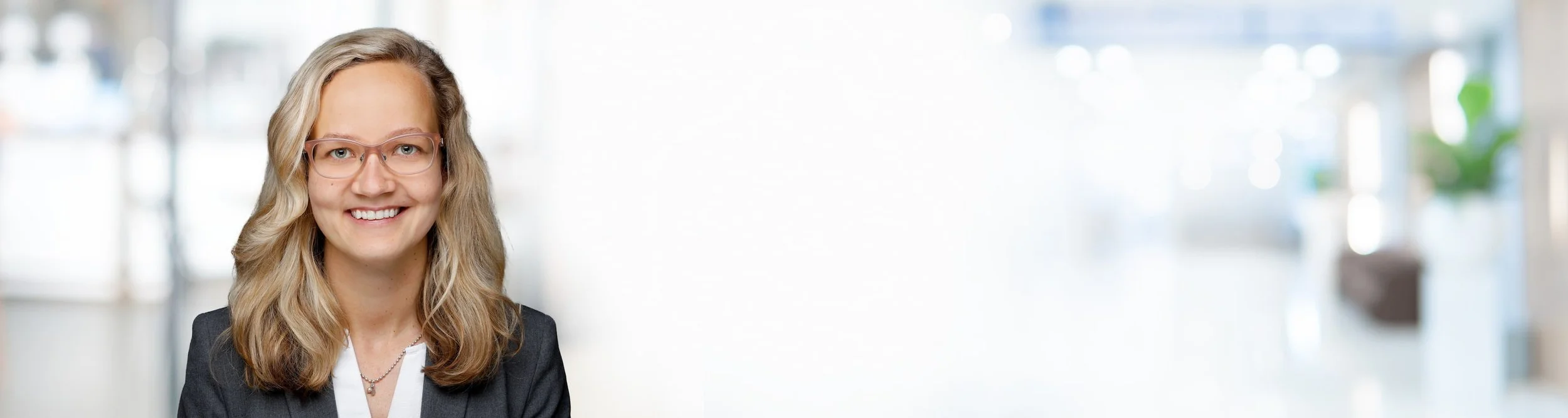 Porträt einer lächelnden Frau mit Brille vor einem hellen, verschwommenen Hintergrund.