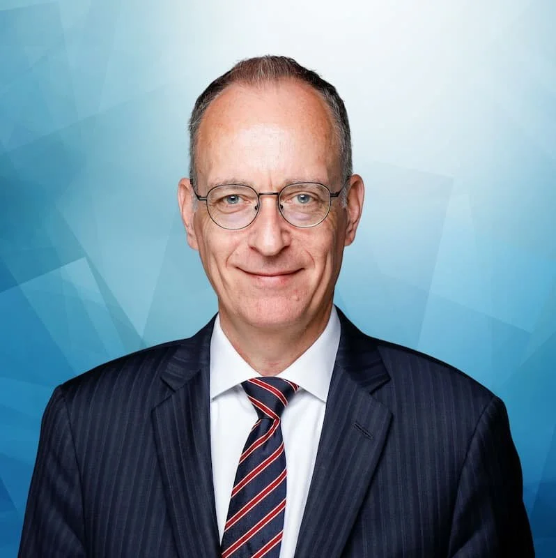 Porträt eines älteren Mannes im Anzug mit Krawatte vor blau-grauem geometrischem Hintergrund.