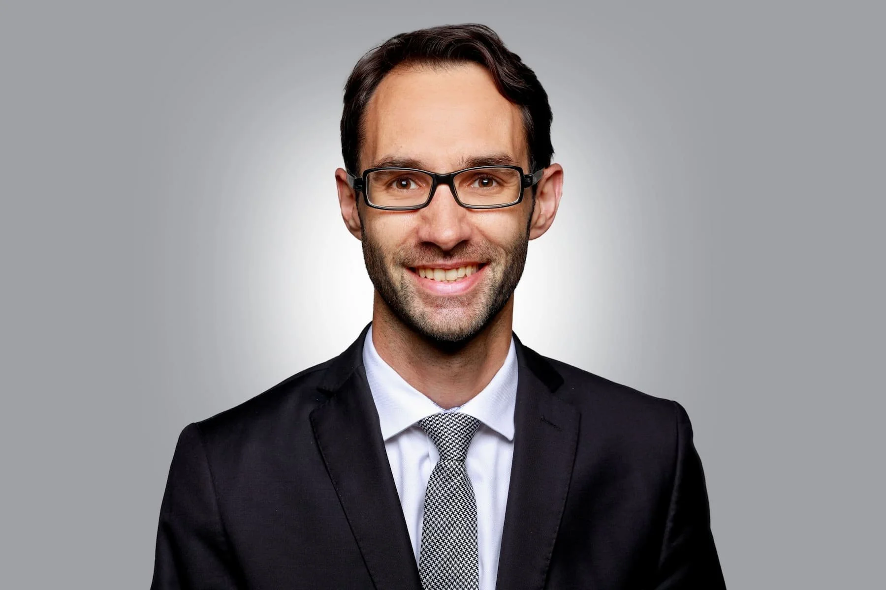 Porträt eines Mannes im Anzug mit Krawatte und Brille, freundlich lächelnd vor neutralgrauem Hintergrund.