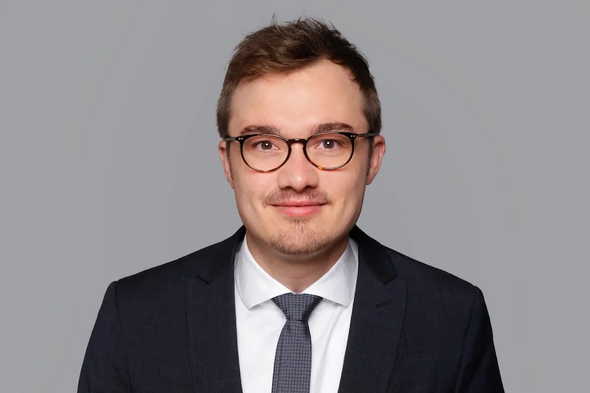 Porträt eines jungen Mannes mit Brille, dunklem Anzug, weißen Hemd und grauem Krawatte vor grauem Hintergrund.