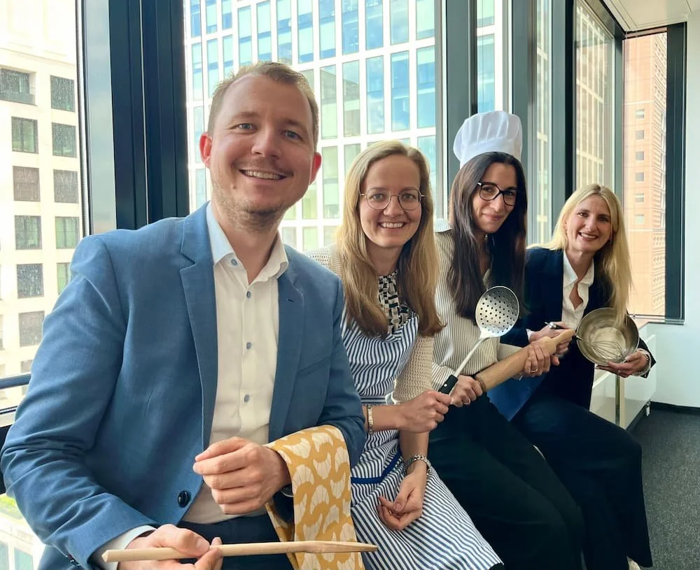 Vier lachende Menschen in Businesskleidung sitzen an einem Fenster in einem Büro. Die Dritte Person trägt eine Kochmütze und hält Kochutensilien, darunter einen Kochlöffel und ein Sieb. Im Hintergrund sind hohe Bürogebäude sichtbar.