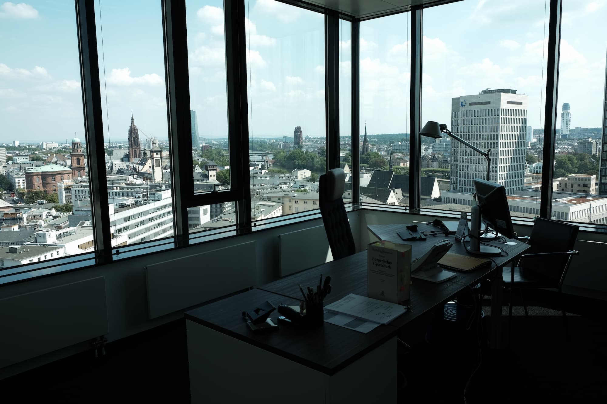 Büro mit großen Fenstern und Stadtblick, Schreibtisch mit Computer, Akten und Büromaterialien.