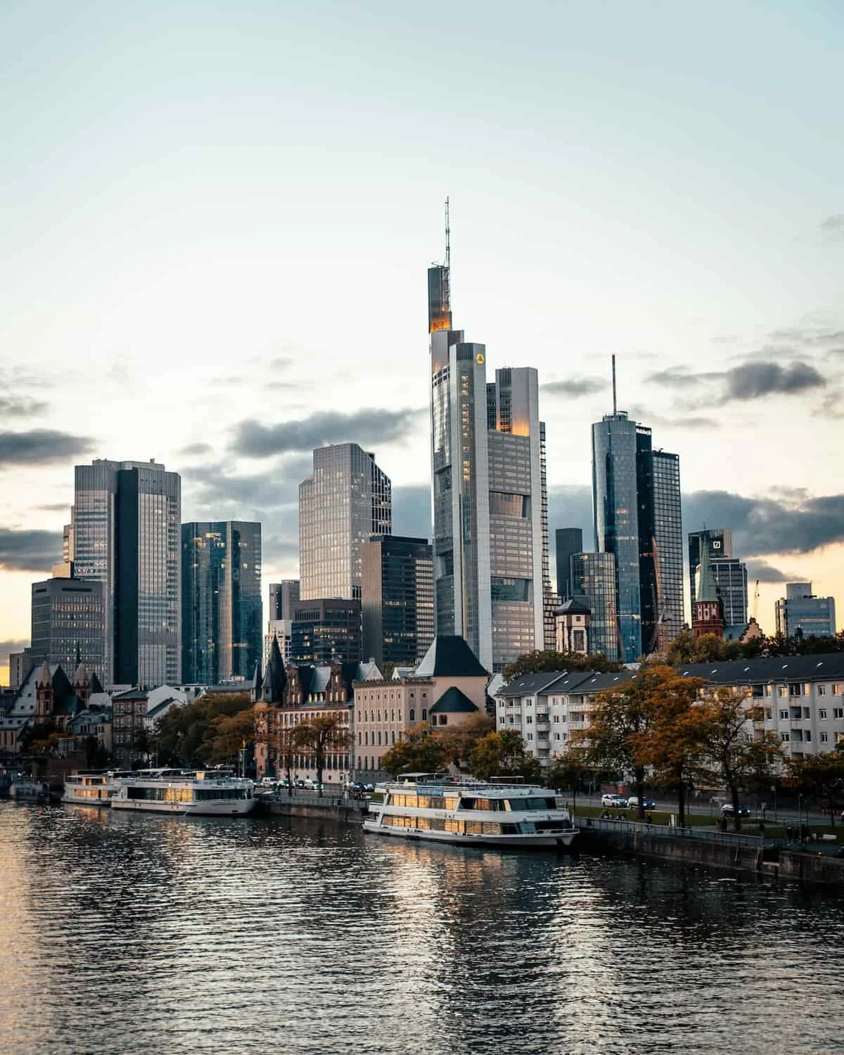 Skyline einer modernen Stadt mit Hochhäusern, Wasser im Vordergrund, Boote und Bäume, Sonnenuntergangsstimmung.
