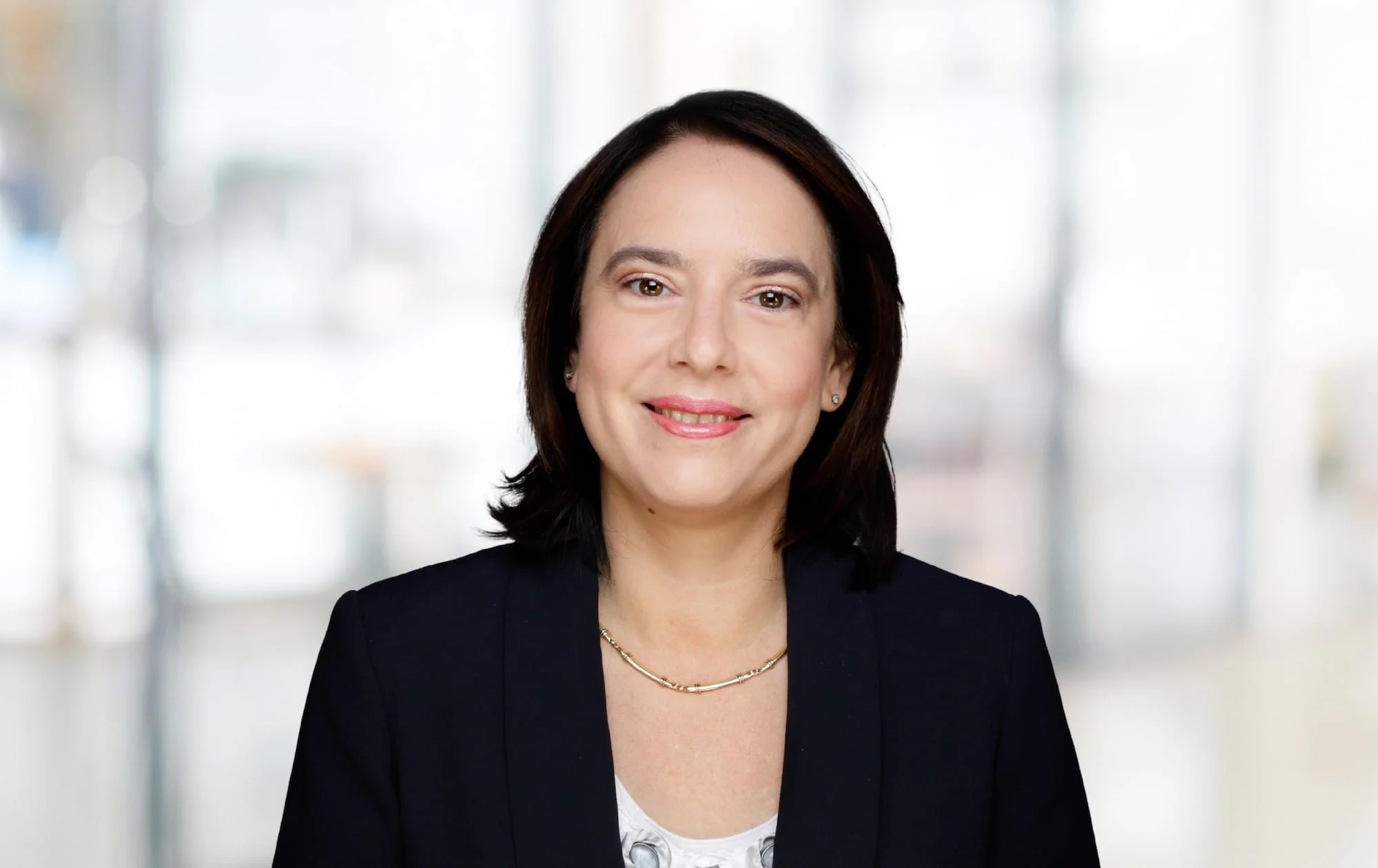 Porträt einer smilingn Frau in dunkler Jacke vor einem hellen, unscharfen Hintergrund.