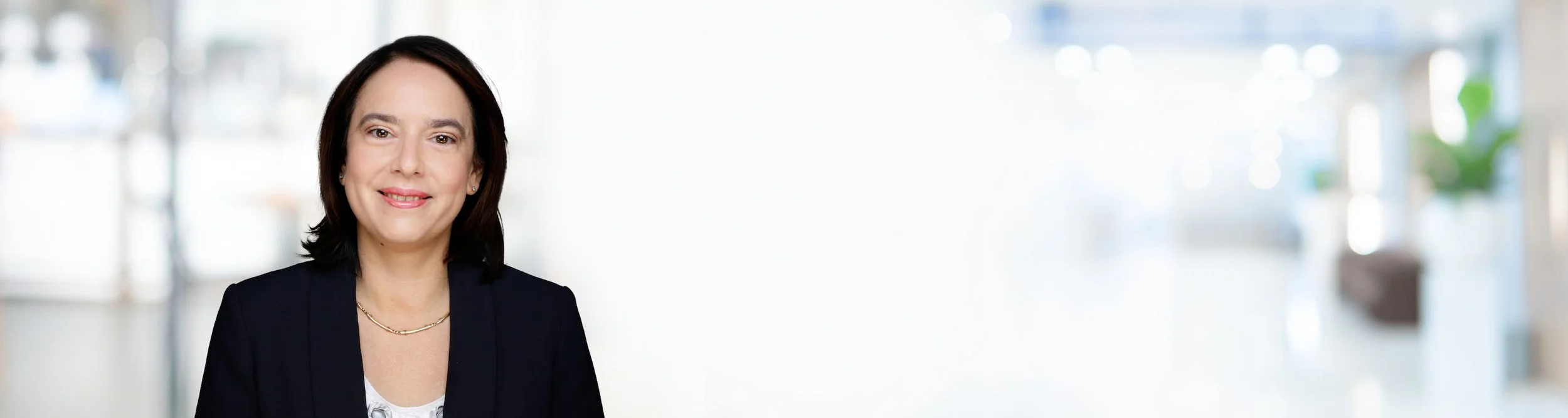 Eine Porträtaufnahme einer smiling Frau mit dunklen Haaren in einem dunklen Blazer vor einem hellen, unscharfen Gebäude im Hintergrund.