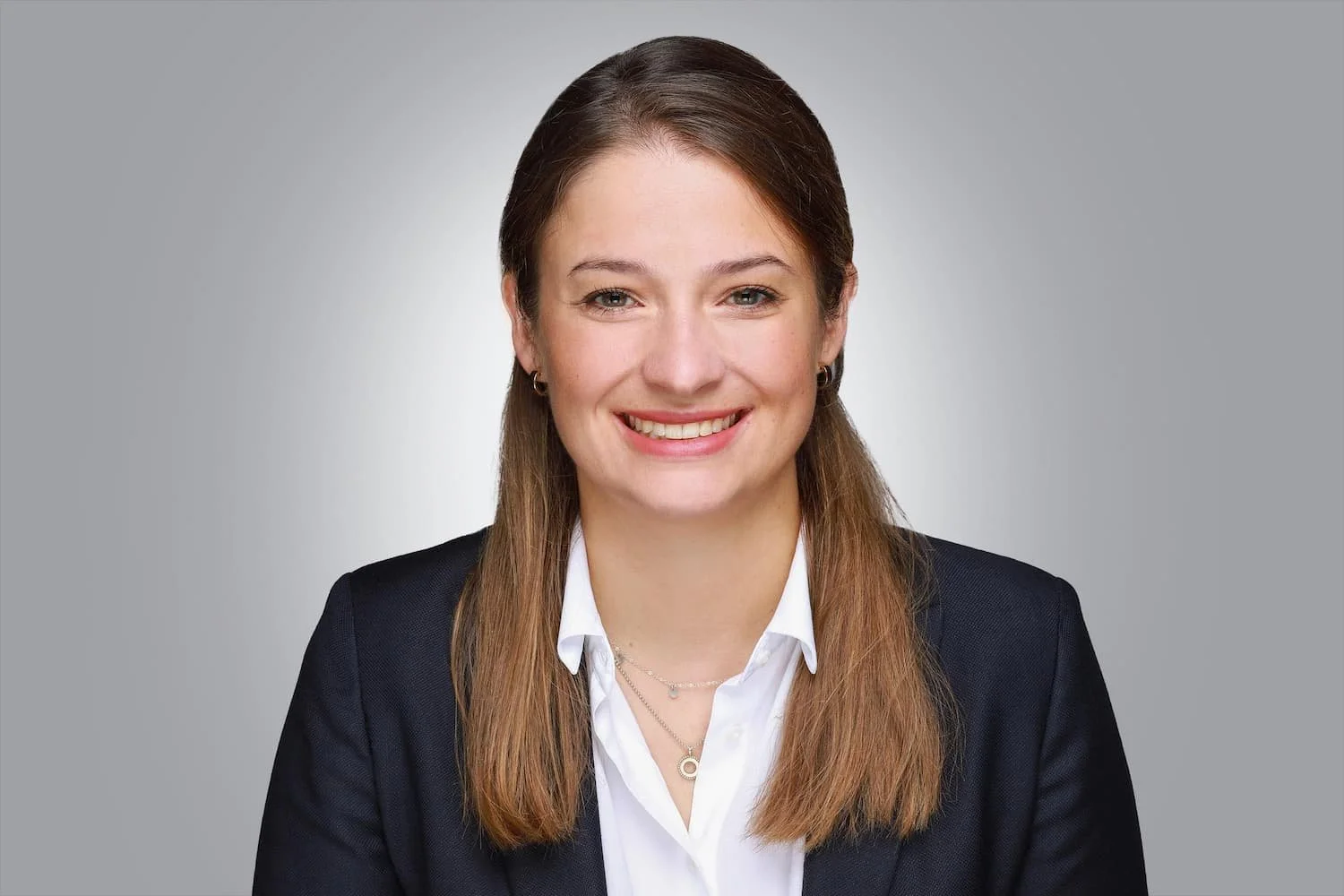 Porträt einer jungen Frau mit braunen Haaren, weißem Hemd und dunklem Blazer vor grauem Hintergrund.