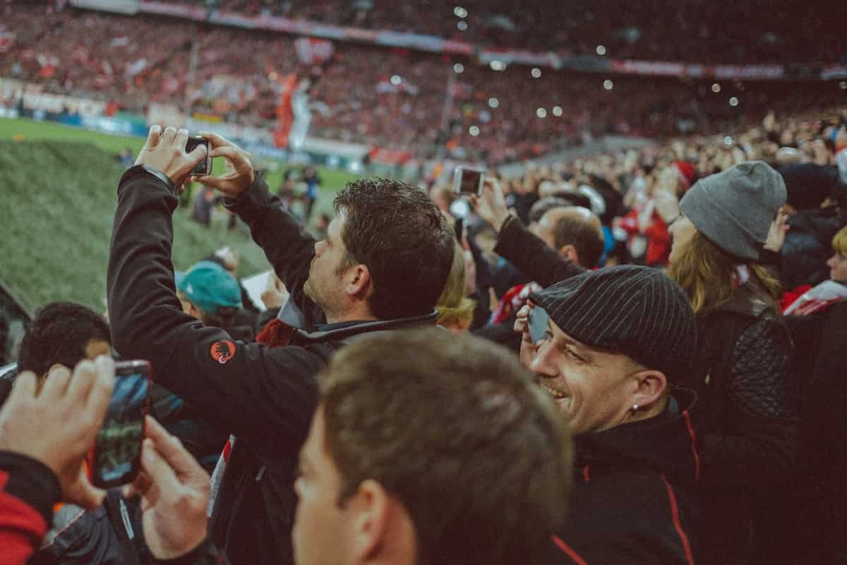 Menschenmenge in einem Stadion, viele Zuschauer fotografieren oder filmen das Spielfeld mit ihren Smartphones, einige tragen Mützen oder Fanartikel.