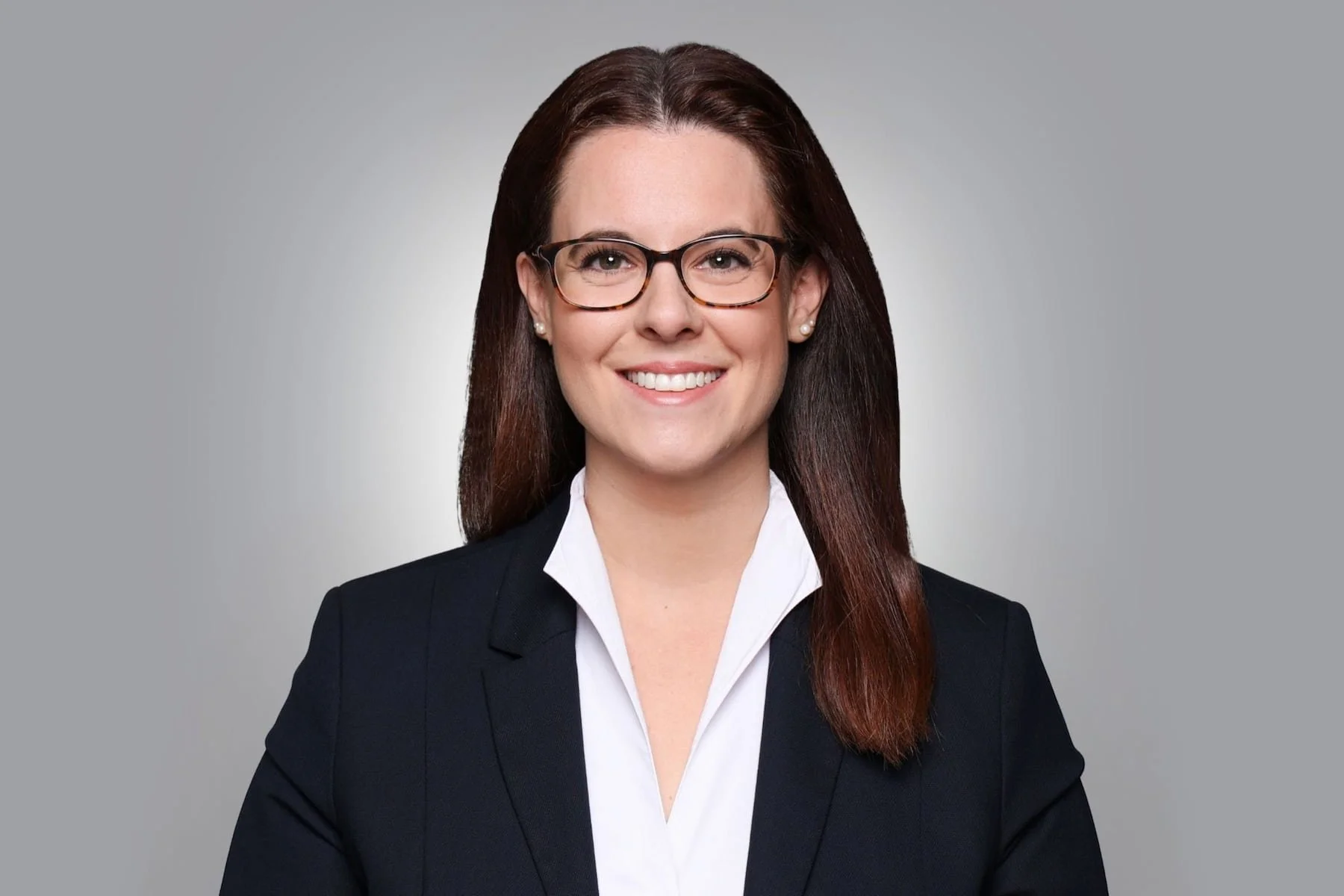 Porträt einer lächelnden Frau mit Brille, dunklen Haaren, Business-Outfit vor hellgrauem Hintergrund.