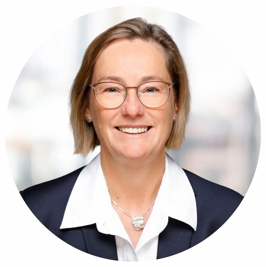 Eine lächelnde Frau mit Brille in einem dunklen Blazer und weißen Hemd vor einem hellen Hintergrund.