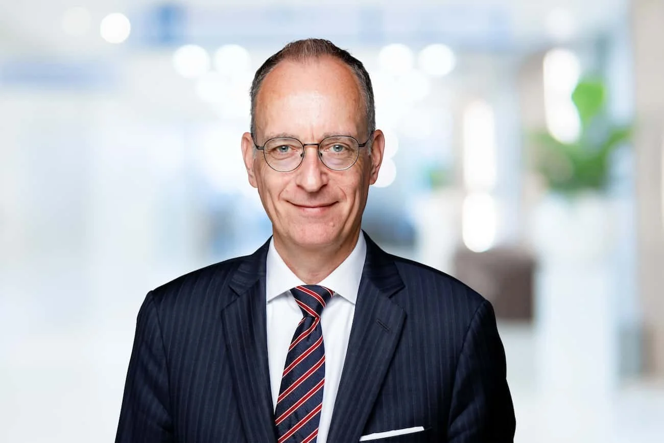 Ein älterer Mann trägt einen dunklen Anzug mit Krawatte und Brille, lächelt und steht in einem lichtdurchflutetem Büro.