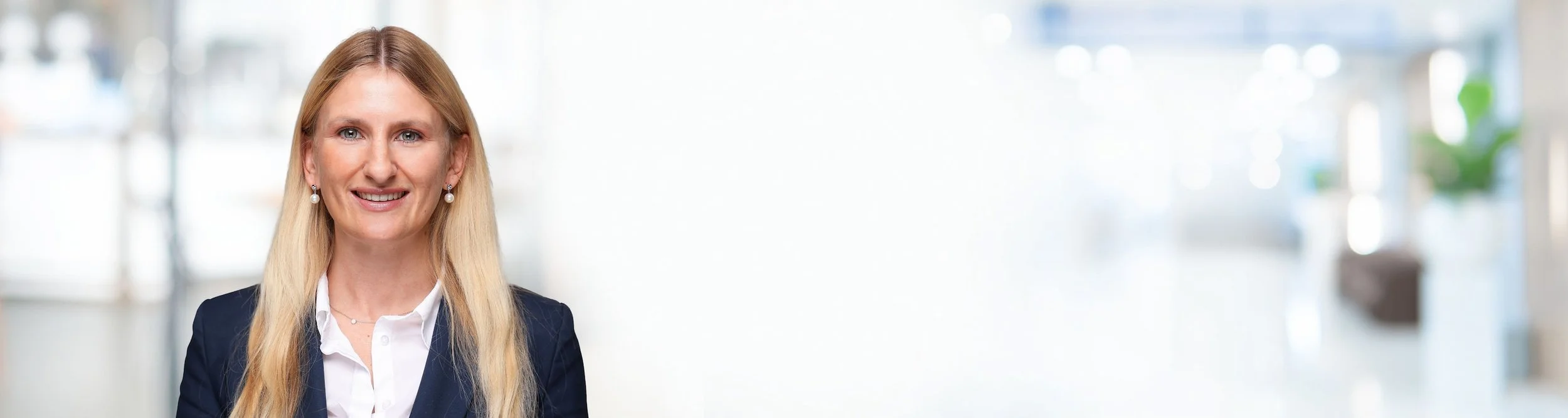 Eine lachende Frau mit blonden Haaren in einem schwarzen Blazer und weißem Hemd steht vor einem hellen, unscharfen Hintergrund mit Fenstern.