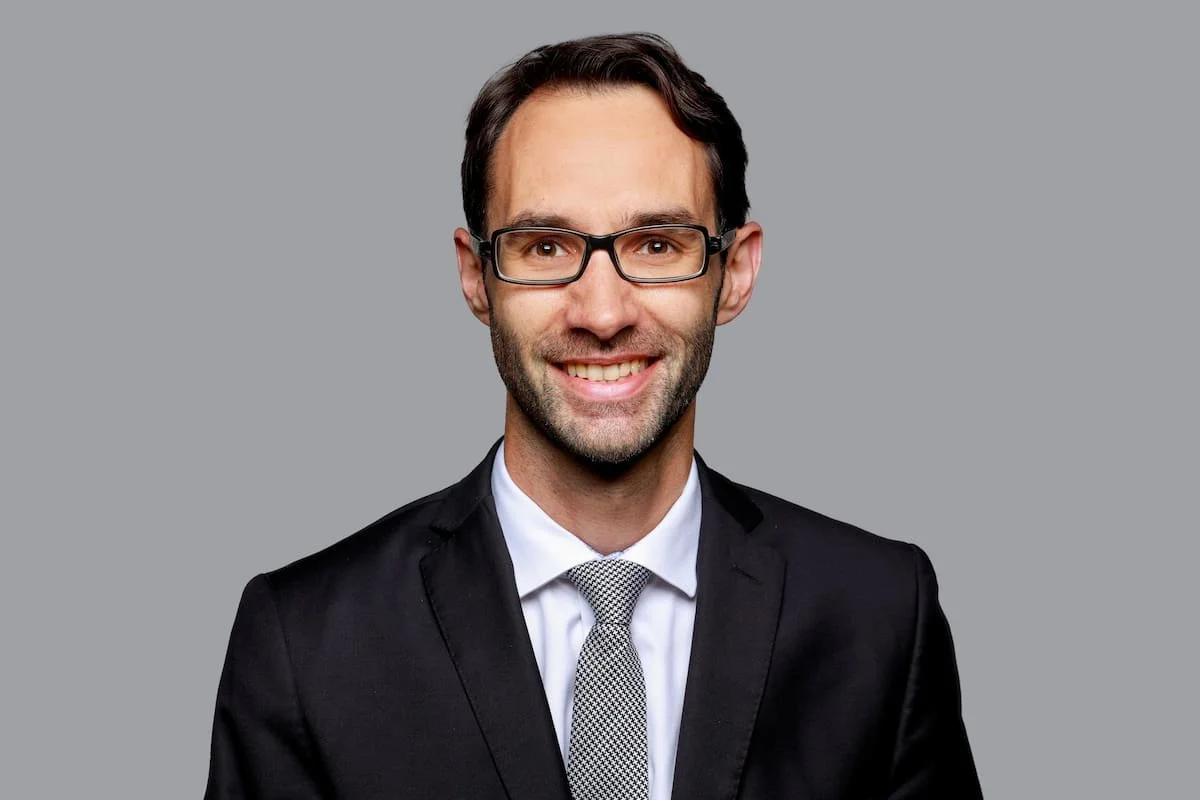 Porträt eines lächelnden Anwalts mit dunklem Anzug, weißem Hemd und grauer Krawatte, Brille, neutraler grauer Hintergrund, professionelles Businessfoto.