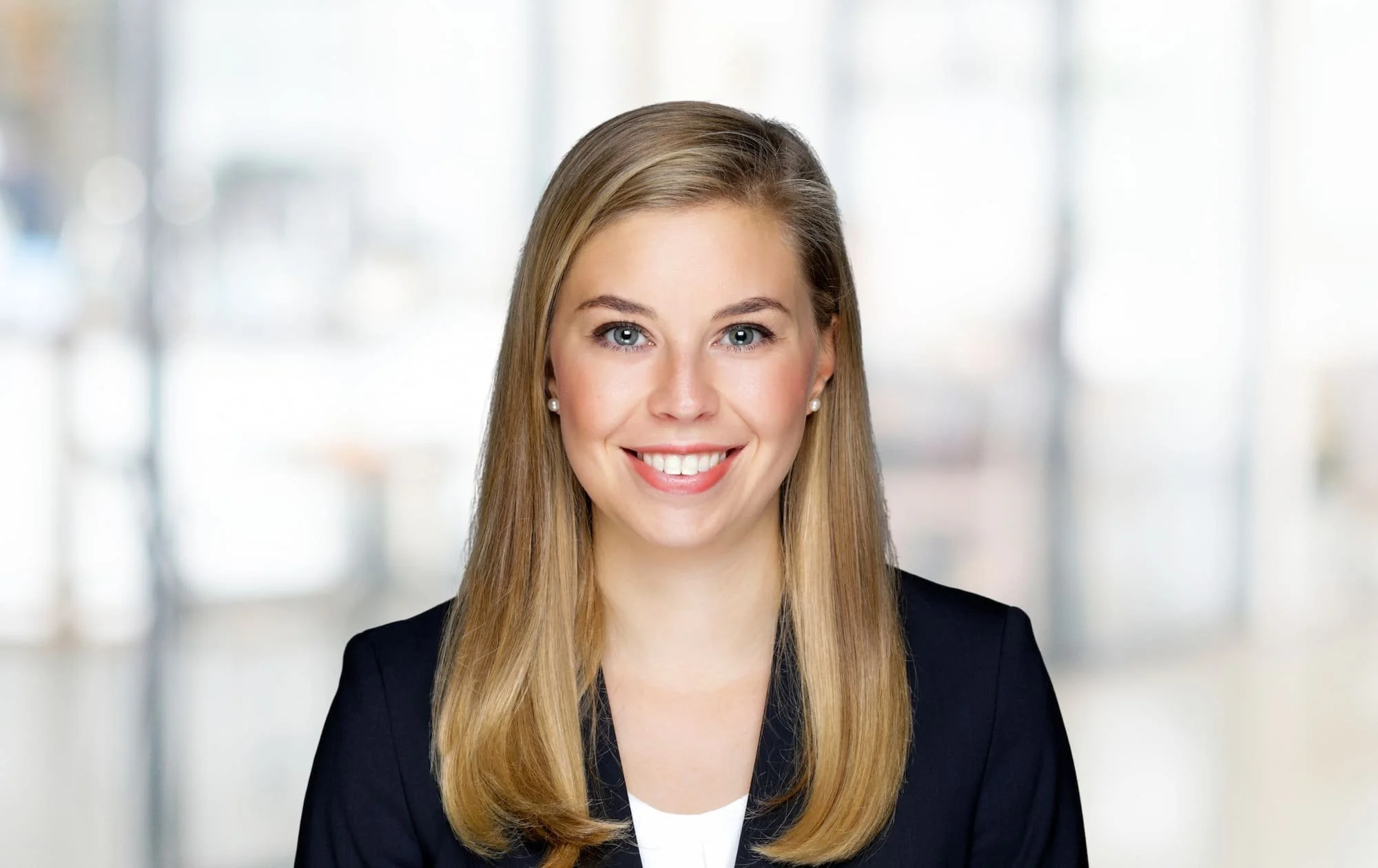 Porträt einer lächelnden Frau in professioneller Kleidung vor unscharfem Hintergrund.