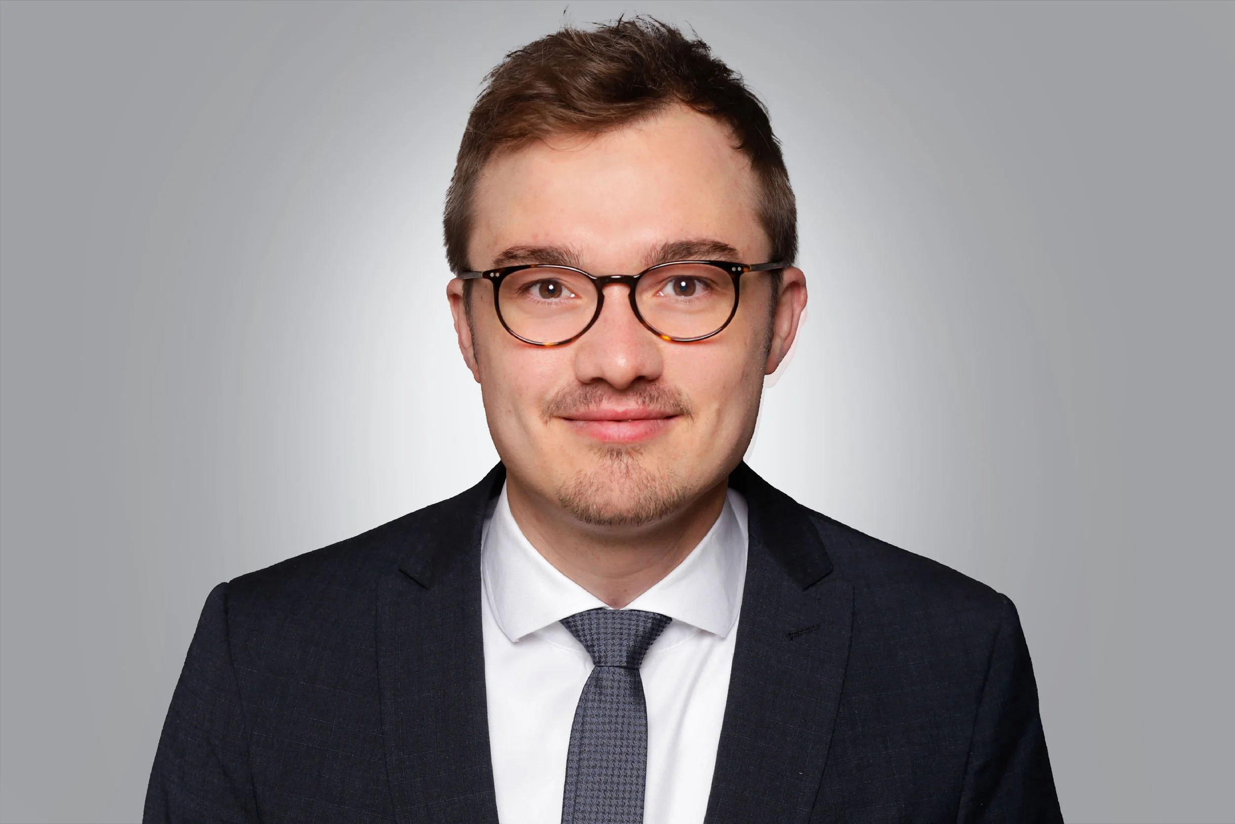 Porträt eines Mannes in Geschäftsanzug, mit Brille, gegen grauen Hintergrund.