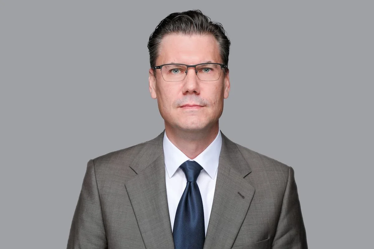 Ein Mann in einem grauen Anzug, weißem Hemd und dunkler Krawatte, mit kurzen, dunklen Haaren und Brille, steht vor einem grauen Hintergrund.