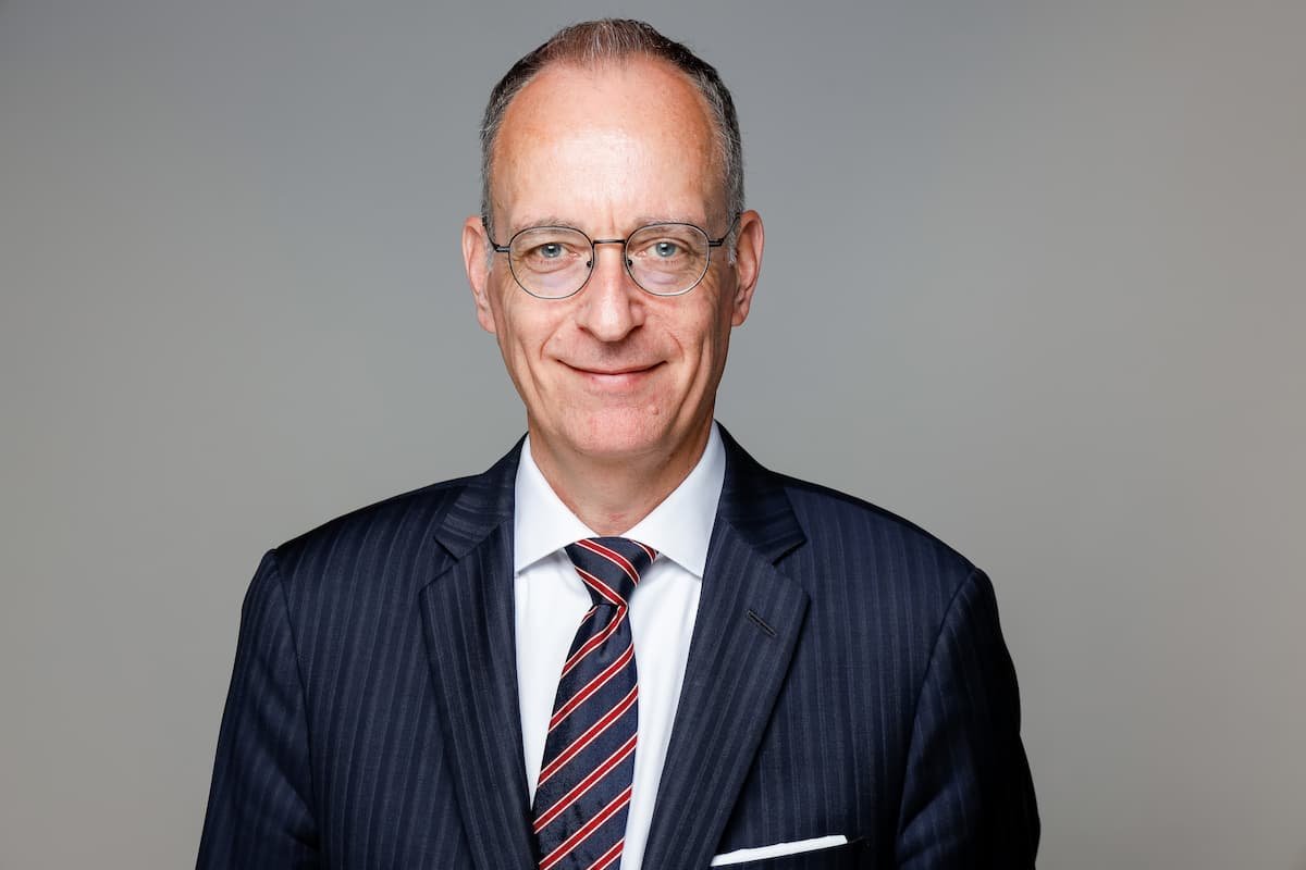 Ein älterer Mann trägt einen dunklen Anzug mit Krawatte und Brille, lächelt leicht, vor einem grauen Hintergrund.