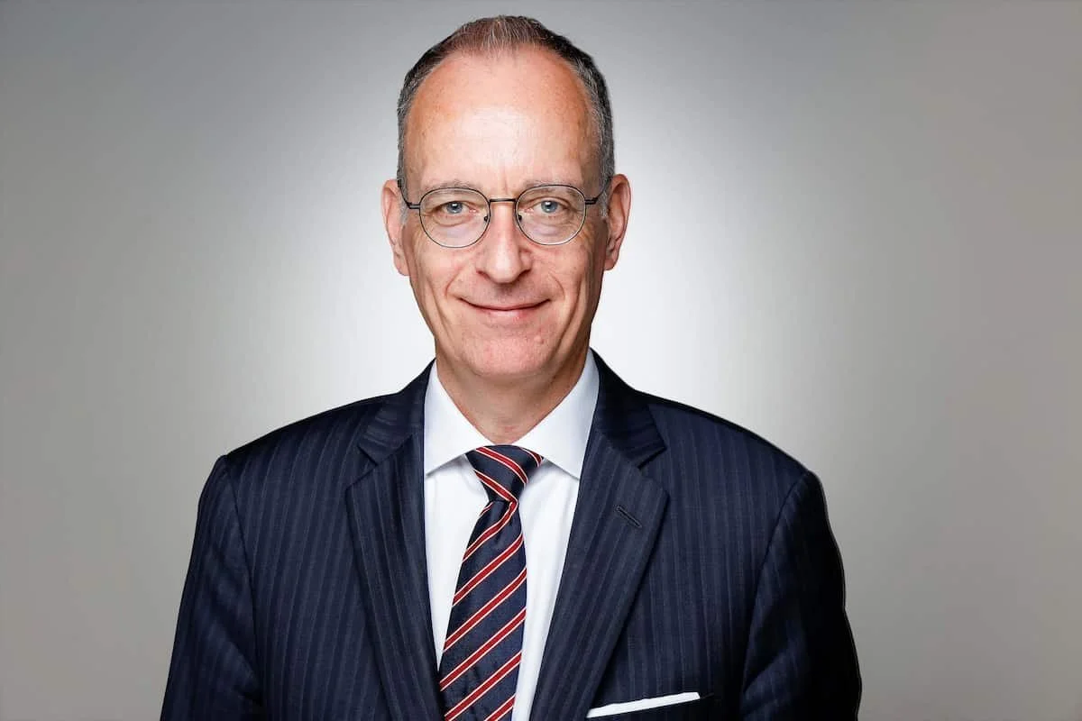 Ein älterer Mann mit freundlichem Gesichtausdruck trägt einen dunklen Anzug mit Krawatte und Brille, lächelt leicht, vor einem grauen Hintergrund.