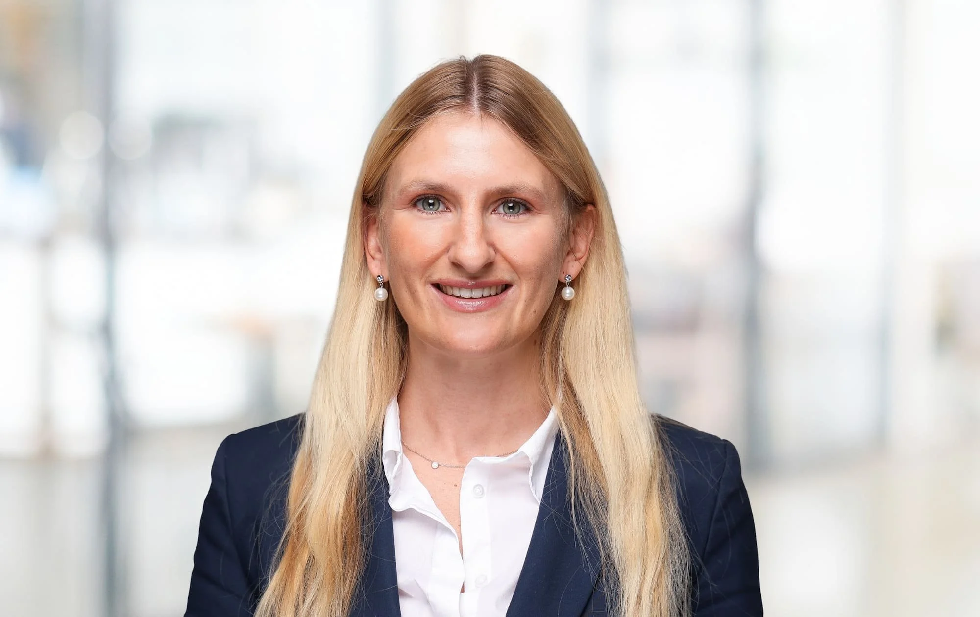 Businessfrau mit langen blondem Haar, dunklem Anzug, weißen Hemd und Perlenohrringen vor einem modernen, hell erleuchteten Hintergrund.