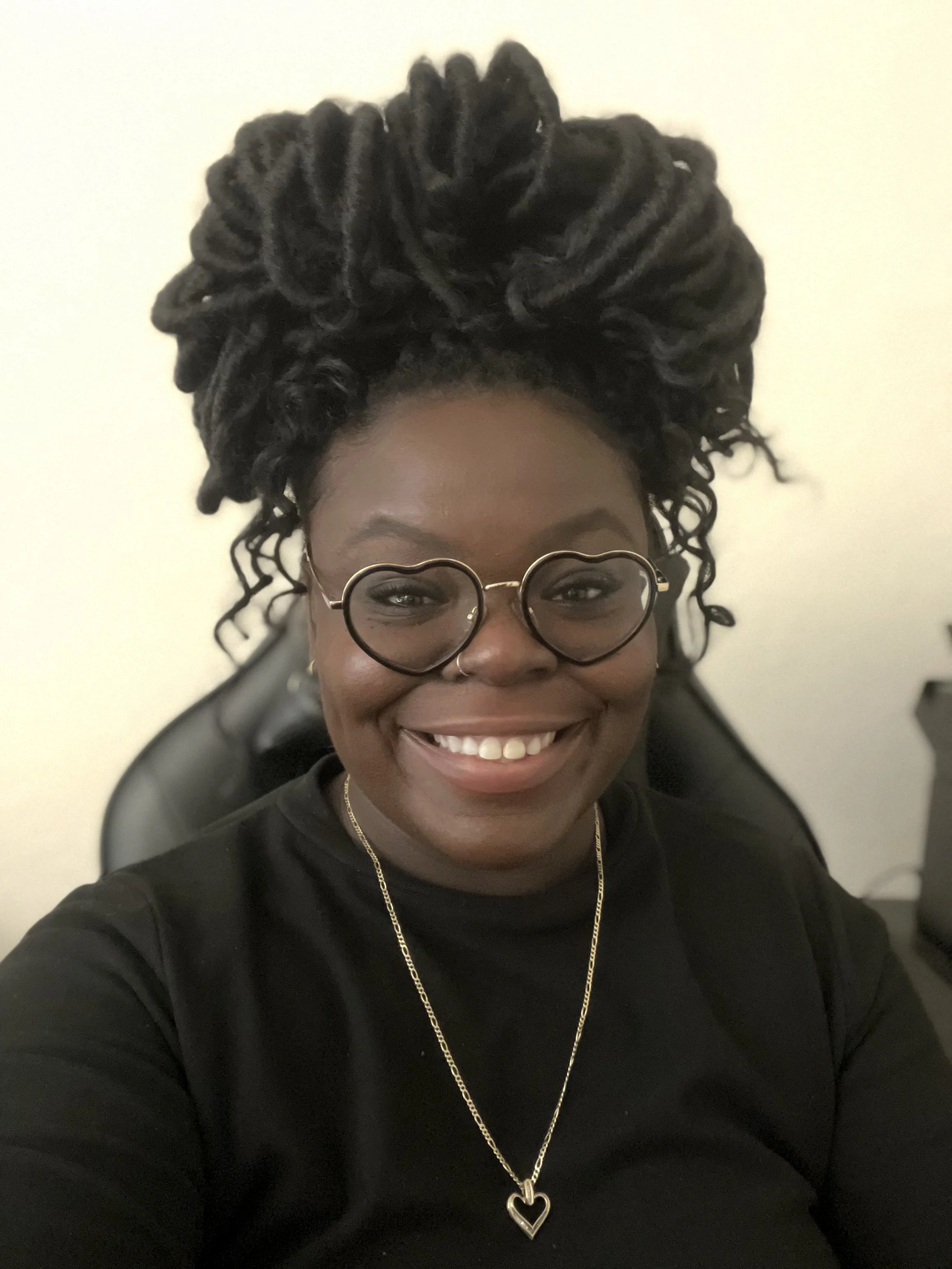 Black woman smiling with heart shaped glasses, a black sweater, and her hair in locs.